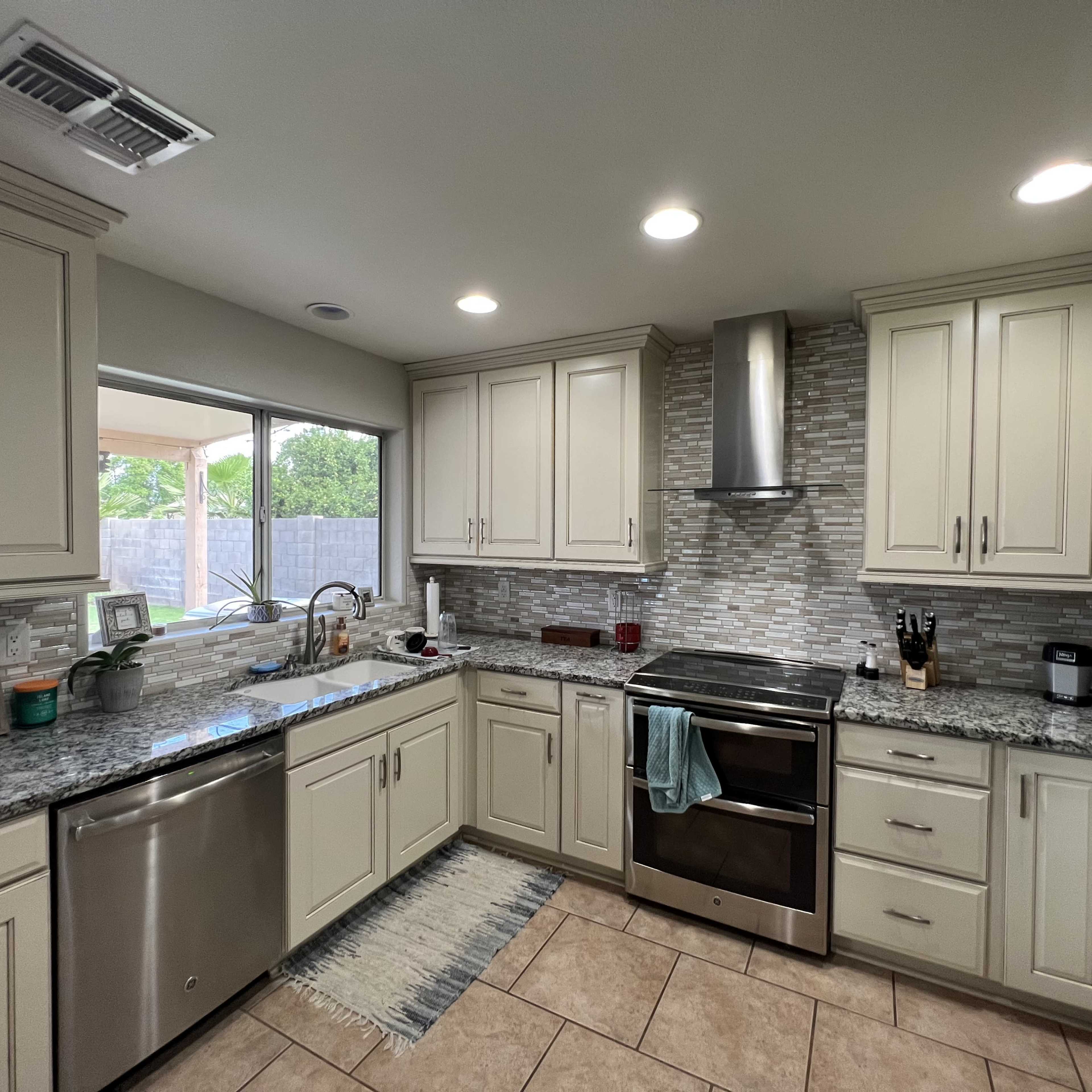 A modern kitchen features beige cabinetry, a stainless steel stove, a stone backsplash, and granite countertops with a window overlooking an outdoor area.