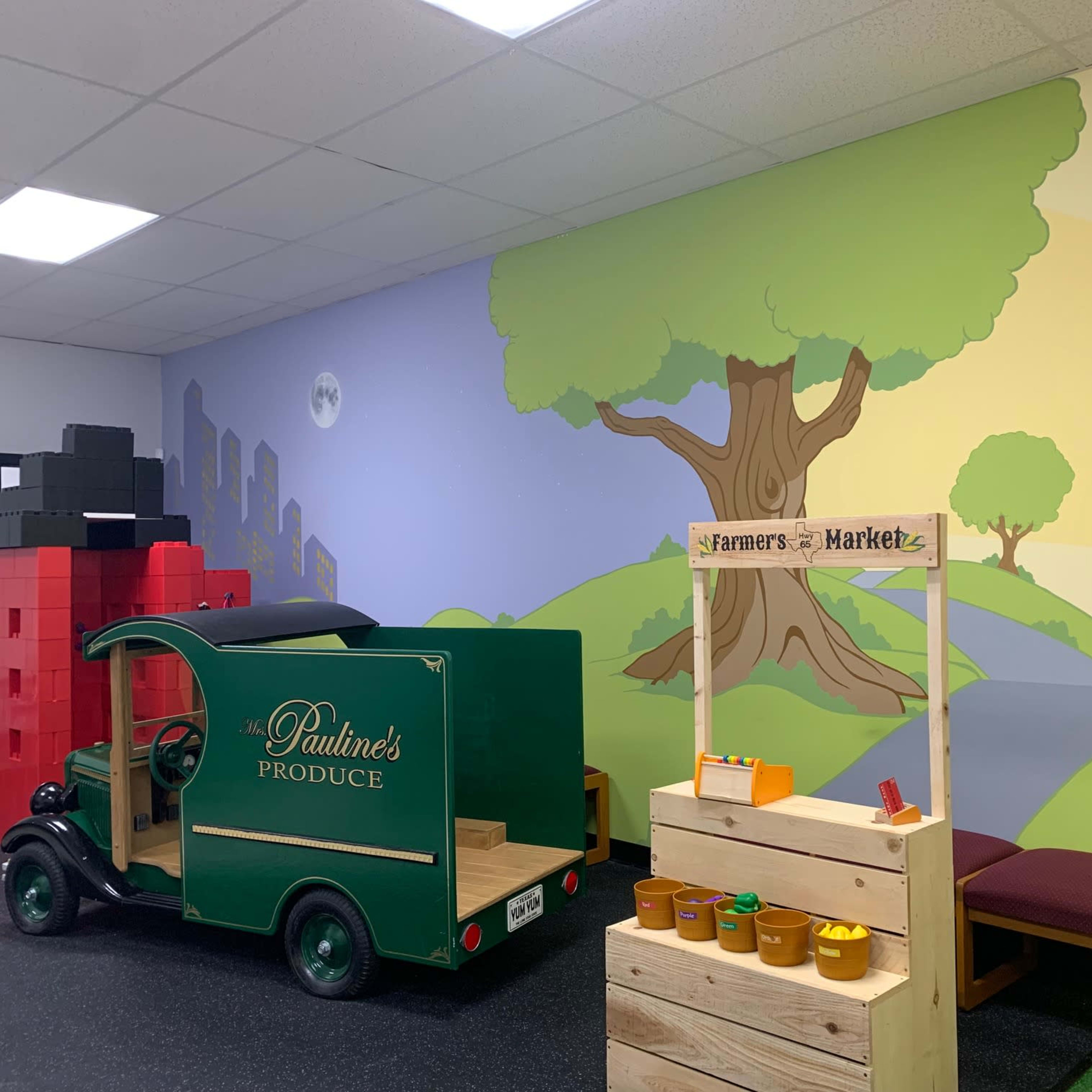 A children's play area featuring a green toy truck labeled "Pauline's Produce," a wooden market stand, and a colorful mural of a tree and a farm market.