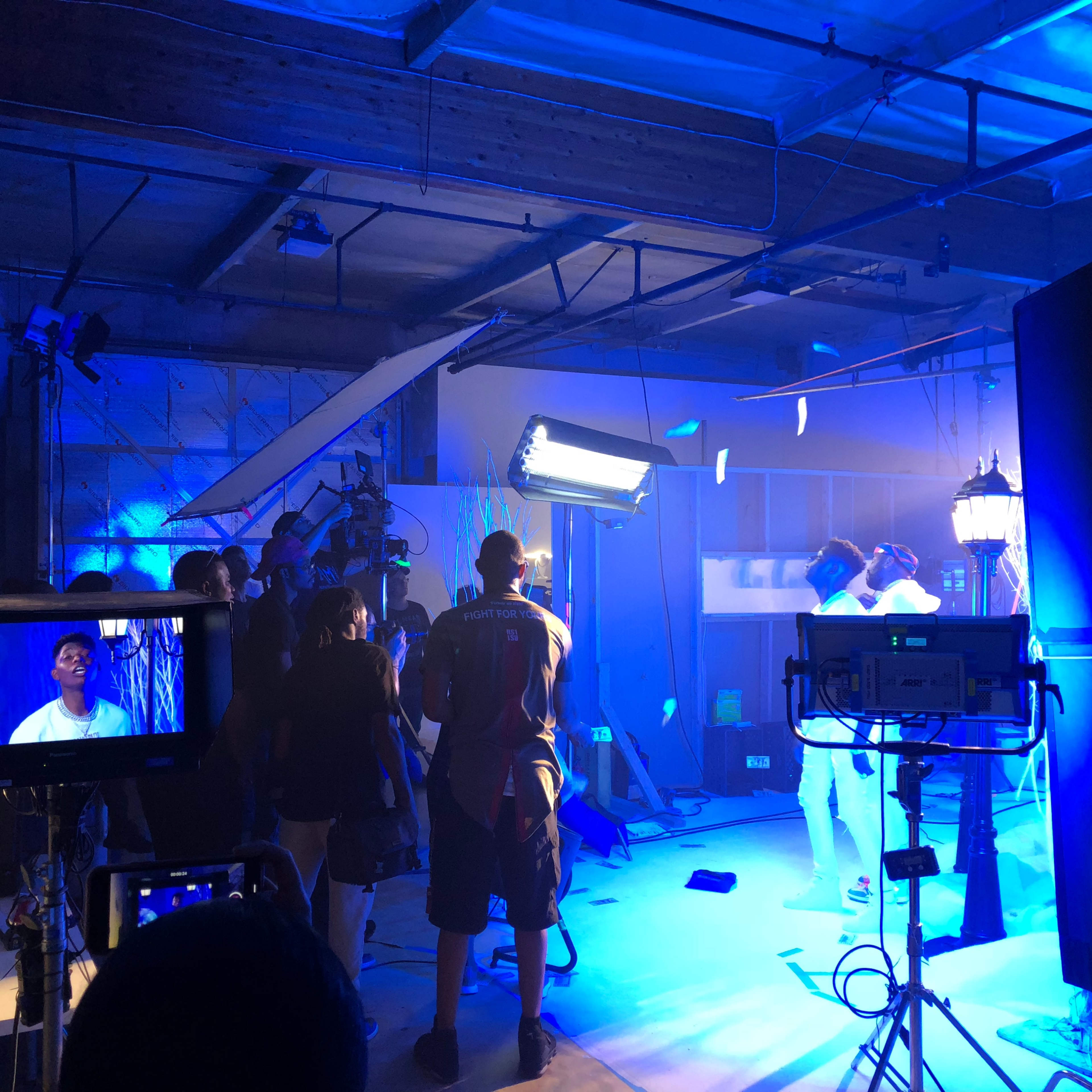 The scene shows a film set with crew members operating cameras and lights in a blue-lit environment.