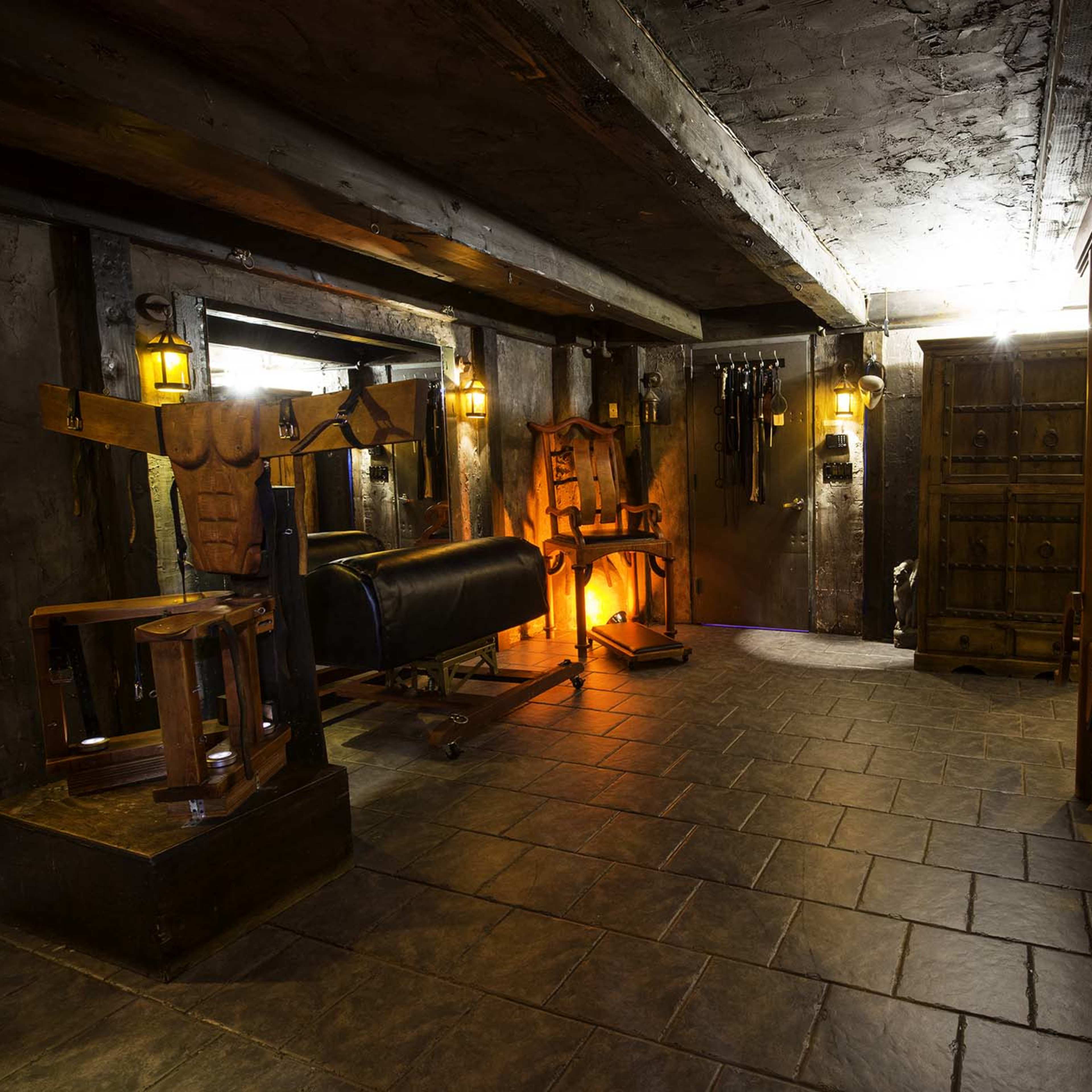 A dimly lit underground room with stone walls, featuring various pieces of exercise equipment and a wooden bench.