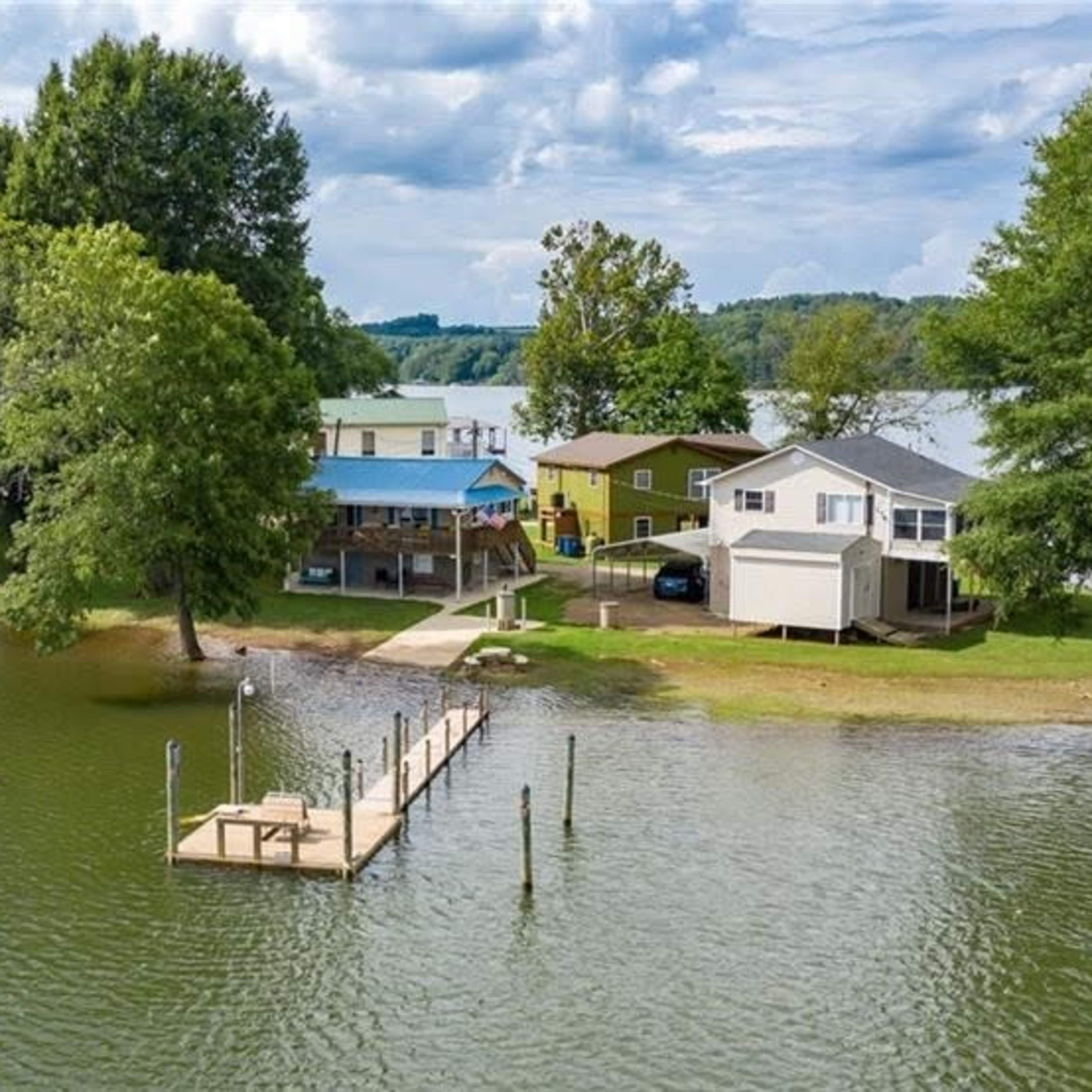 A view of a lakeside property featuring multiple buildings, docks, and lush trees under a partly cloudy sky.