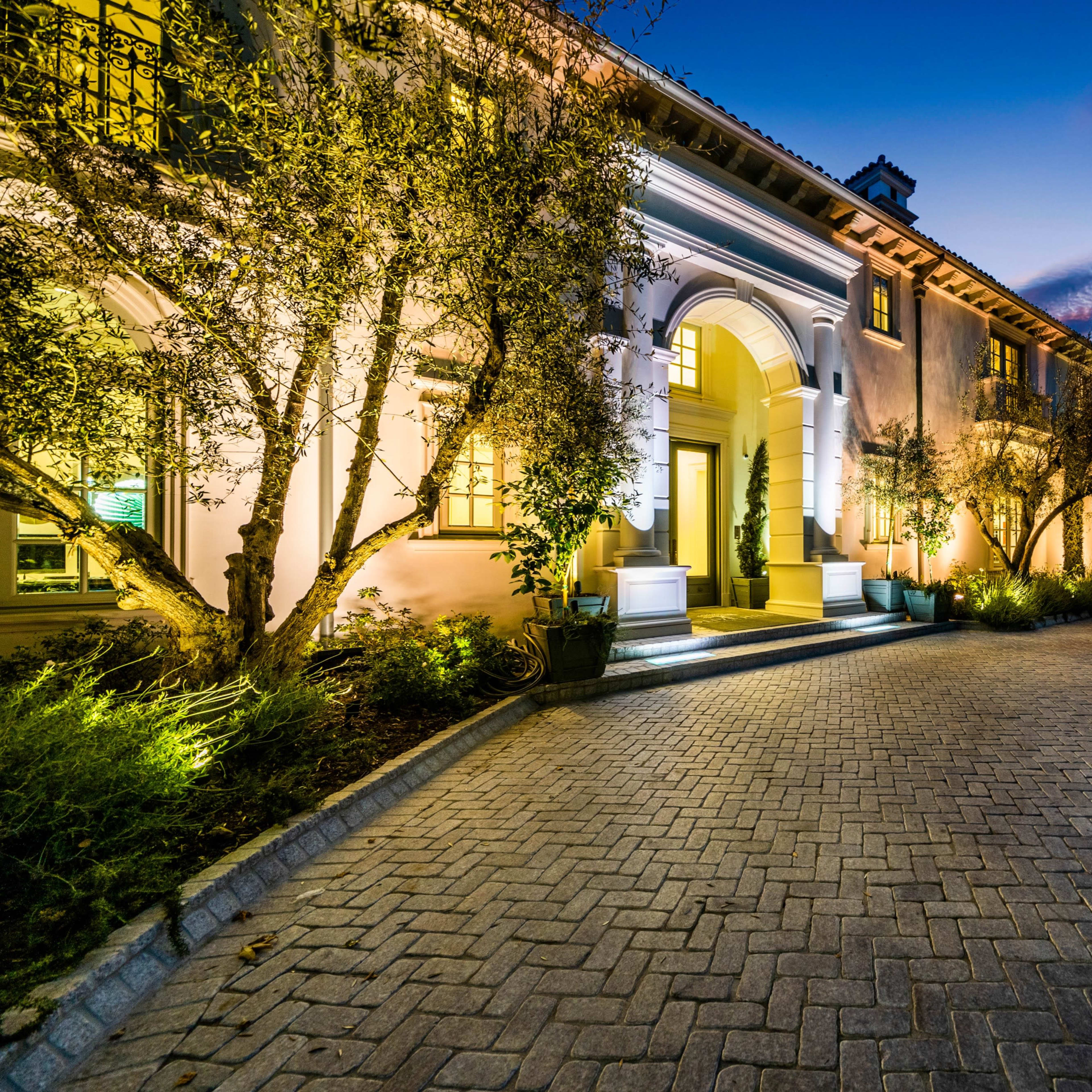 A well-lit luxury home features a winding brick driveway leading to an elegant entrance surrounded by landscaped trees and shrubs.