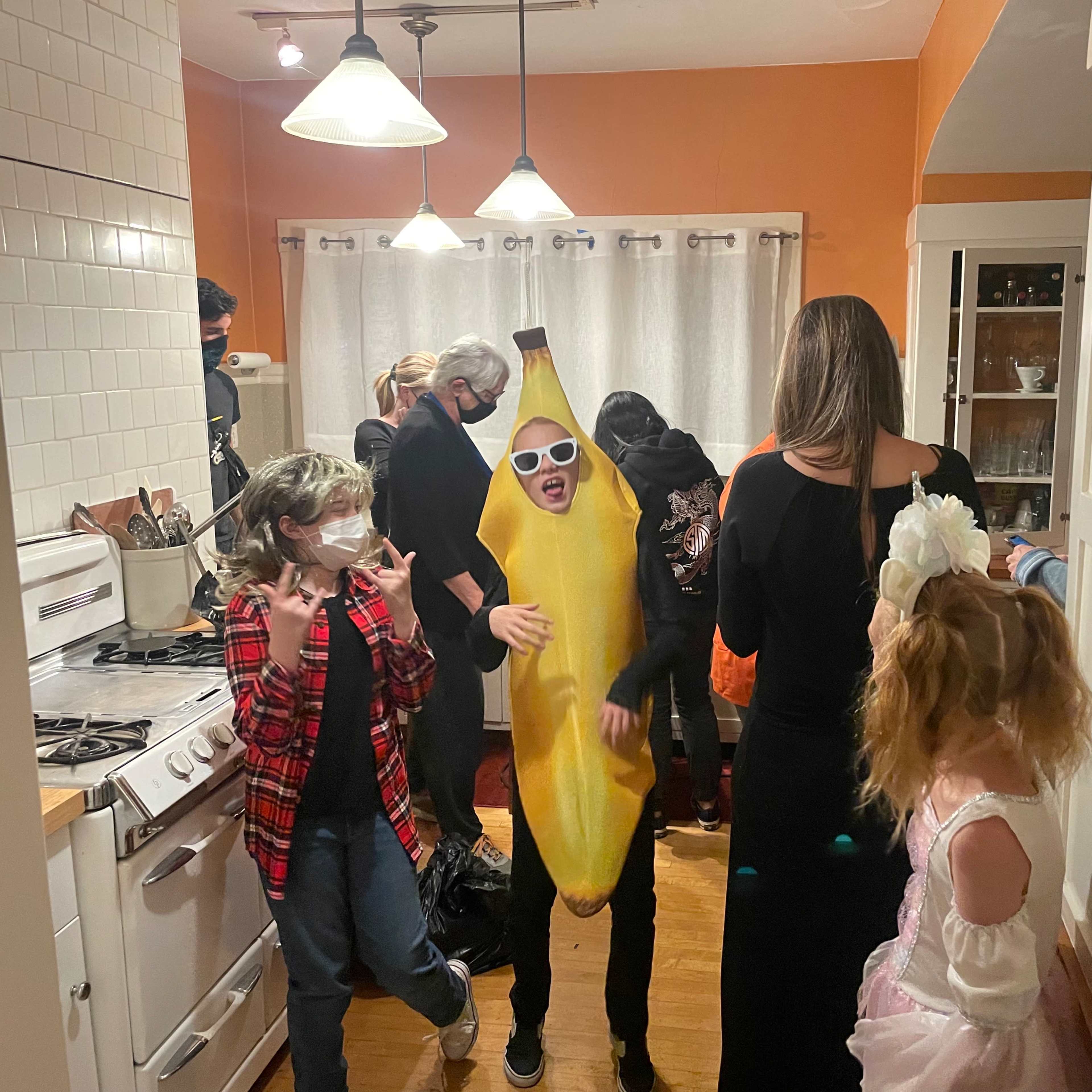 A group of people gathers in a kitchen, with one person dressed in a banana costume and others engaged in conversation and activities.