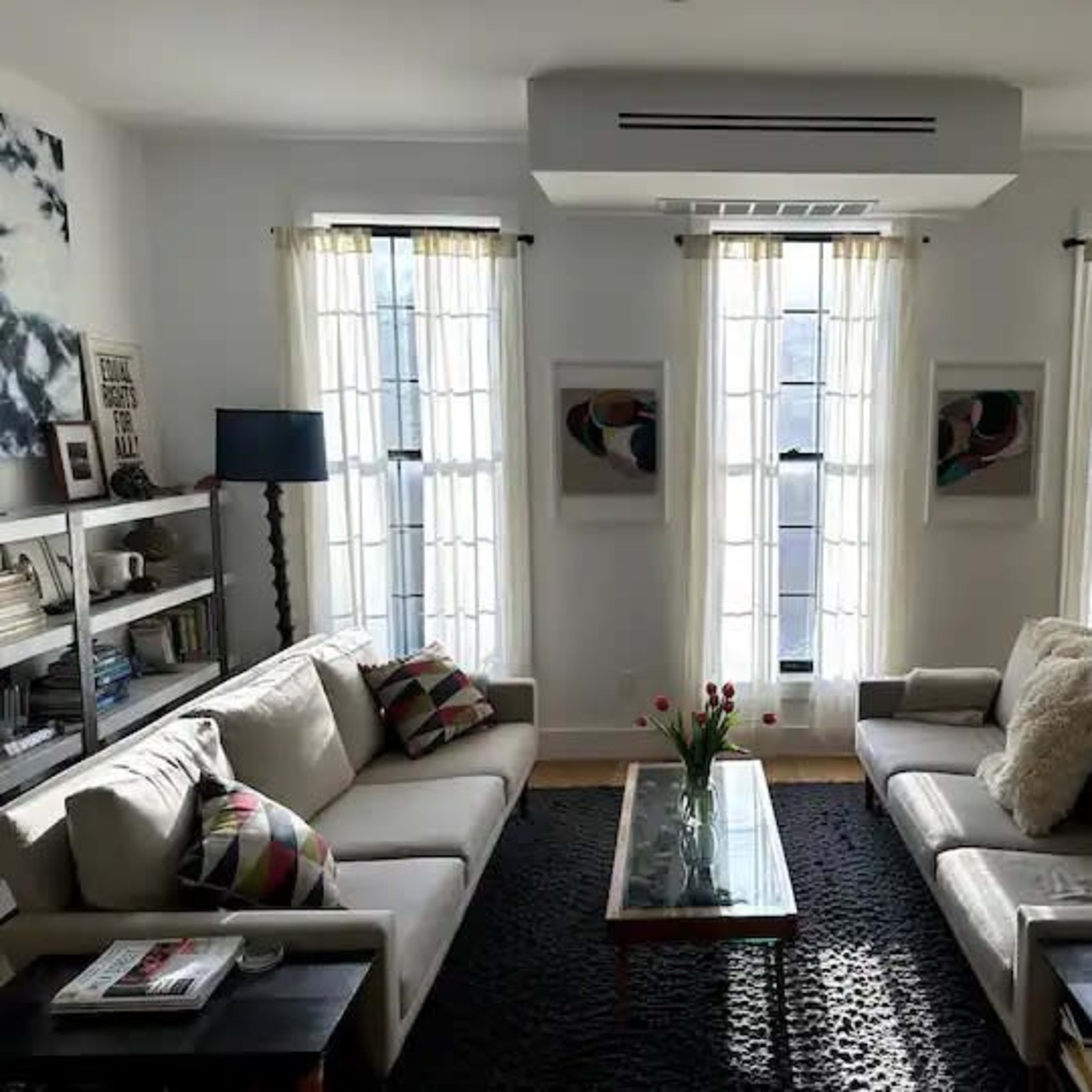 The living room features two sofas arranged around a glass coffee table, with large windows dressed in sheer curtains and a bookshelf along one wall.