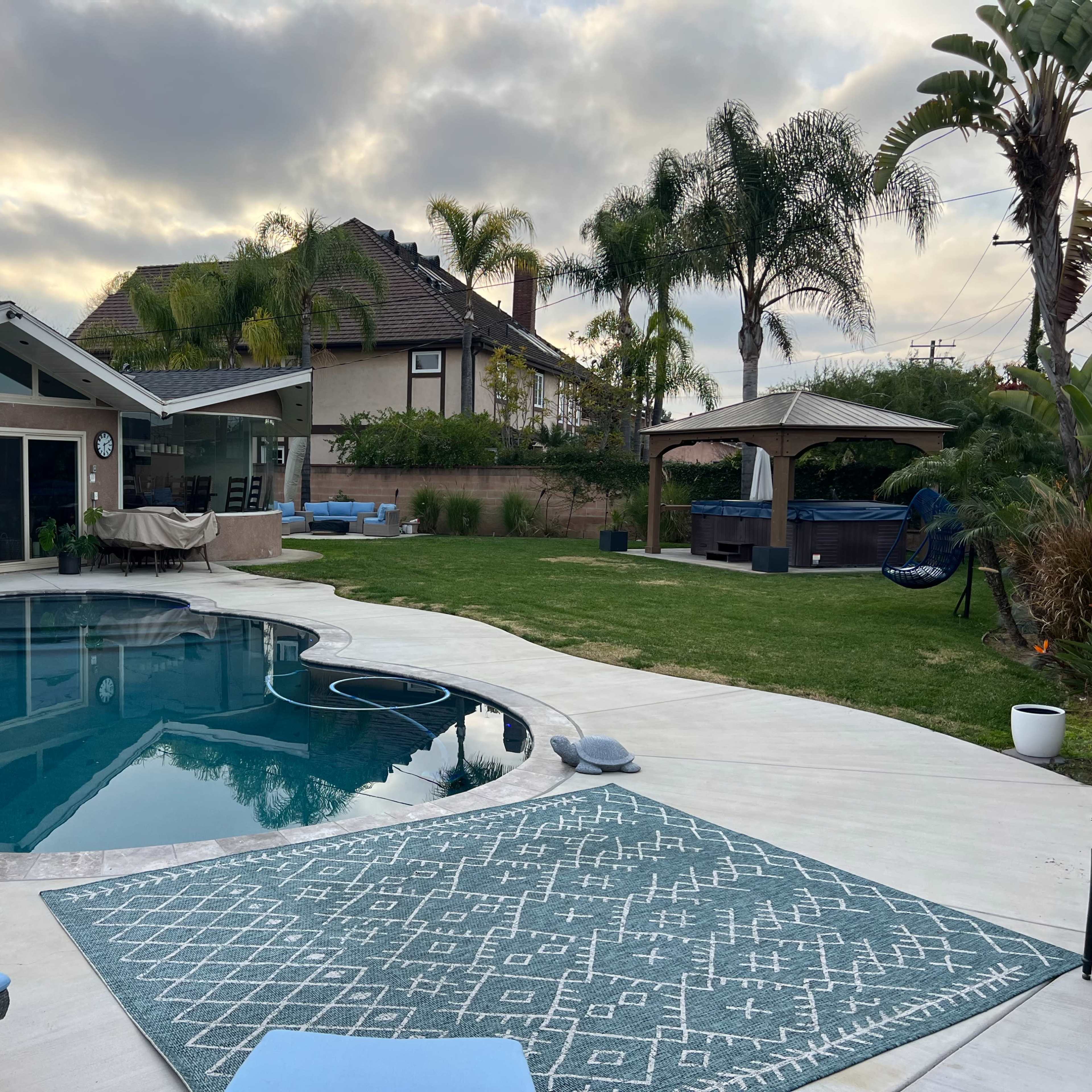 A backyard with a swimming pool, lounge chairs, and a well-maintained lawn surrounded by palm trees and a house.