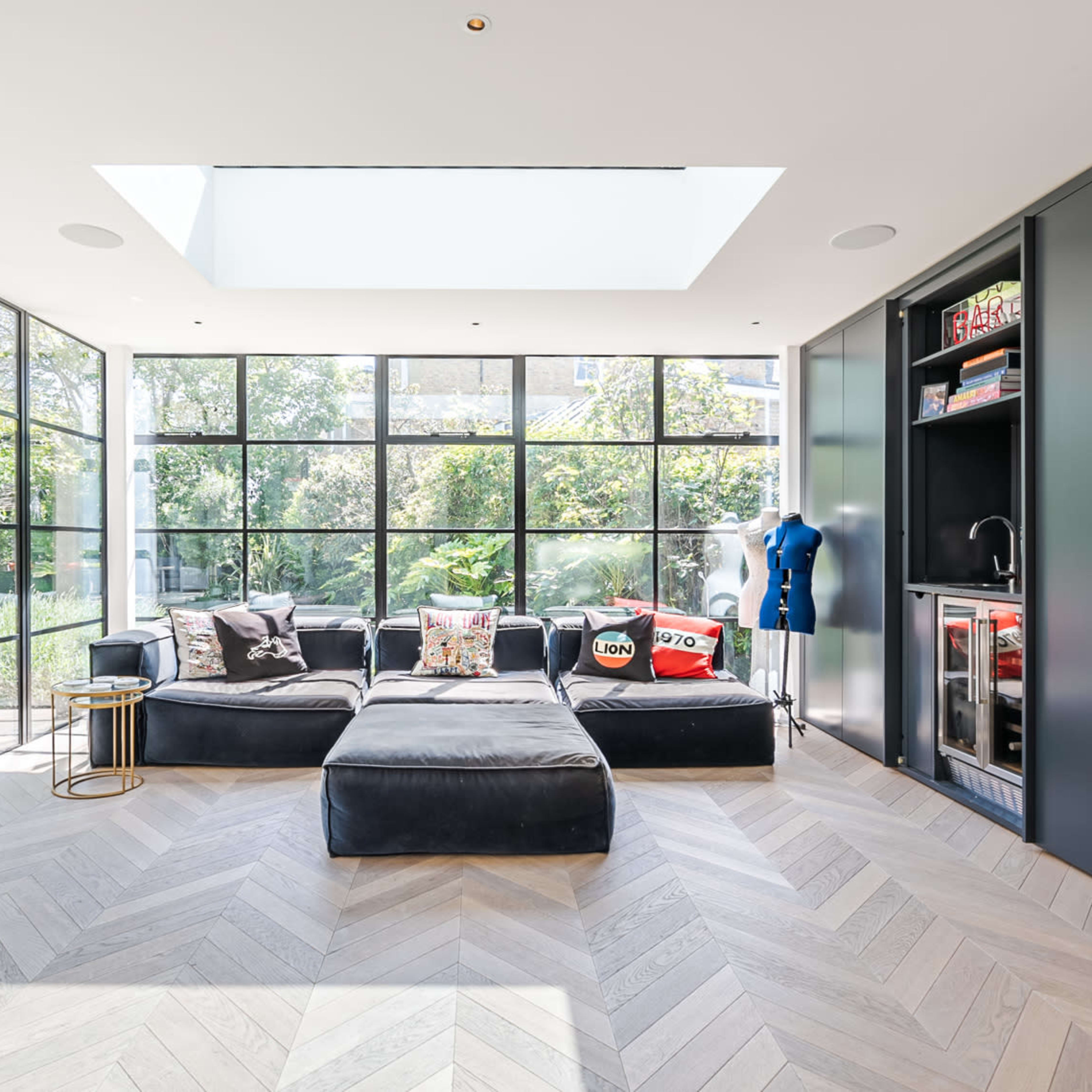 A modern living room features large windows that offer a view of greenery, with a black sectional couch and a small side table.