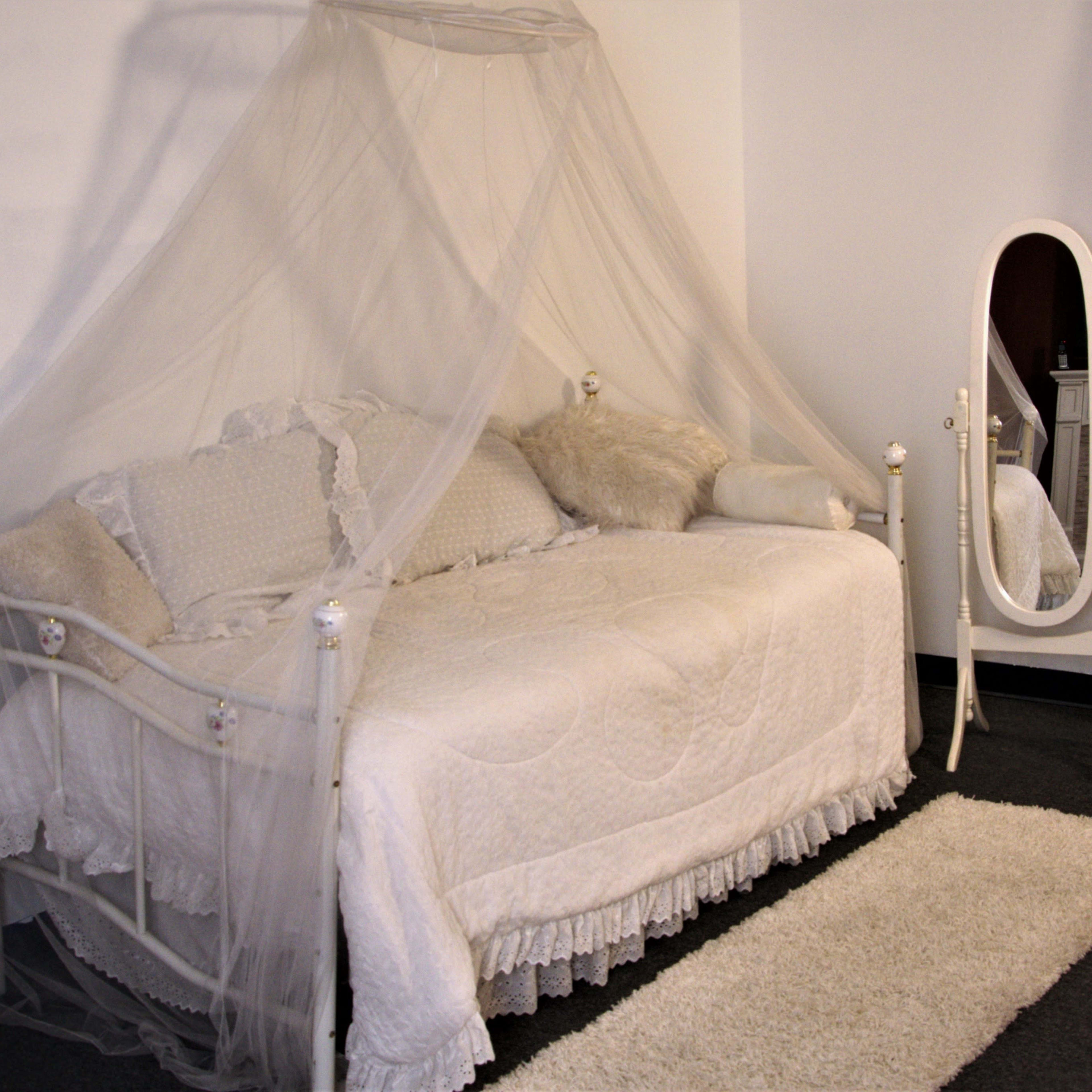 A bedroom with a canopied daybed draped in light fabric next to a full-length mirror and a soft rug on the floor.
