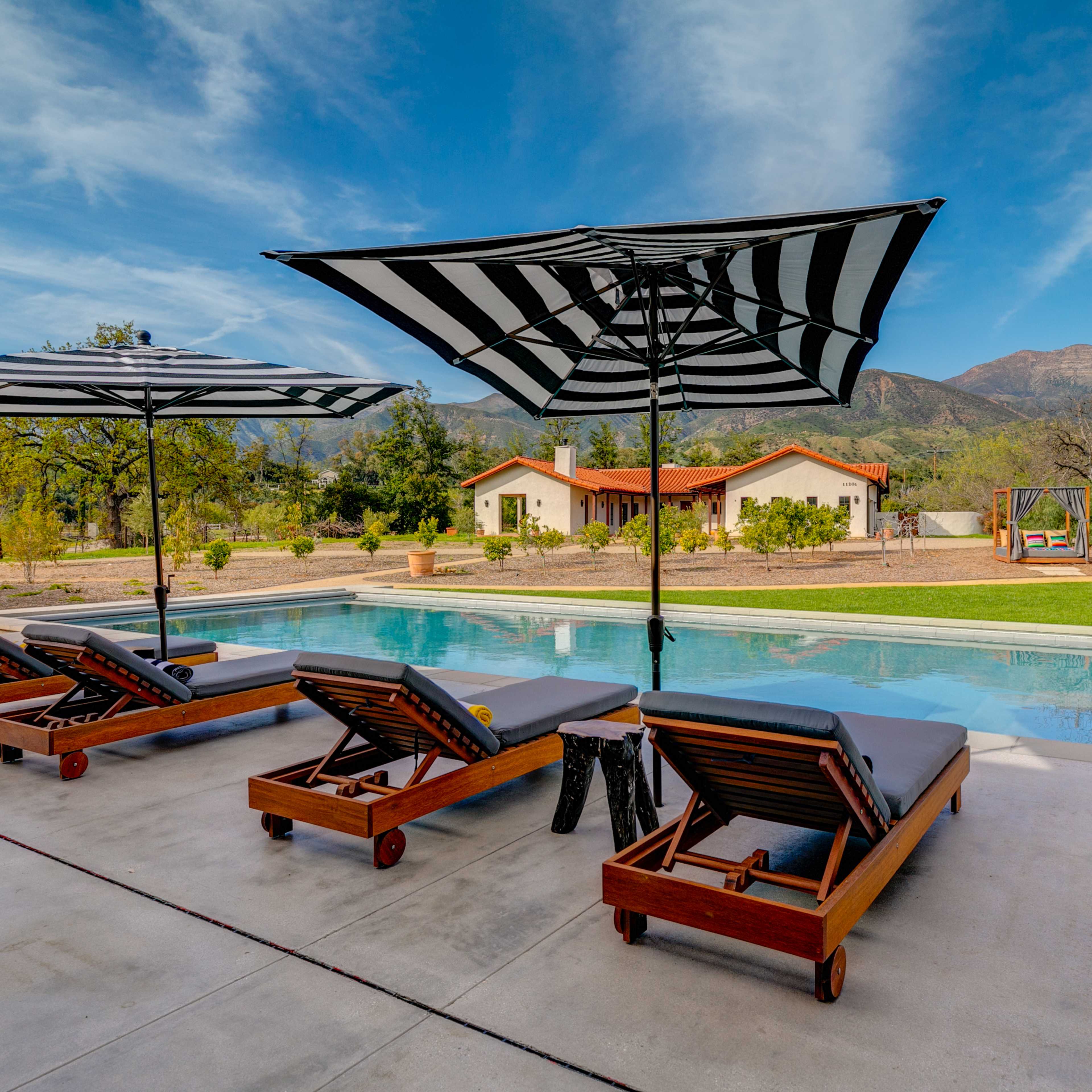 A serene outdoor setting with four loungers under black and white striped umbrellas, next to a swimming pool and surrounded by a landscaped garden and mountains in the background.