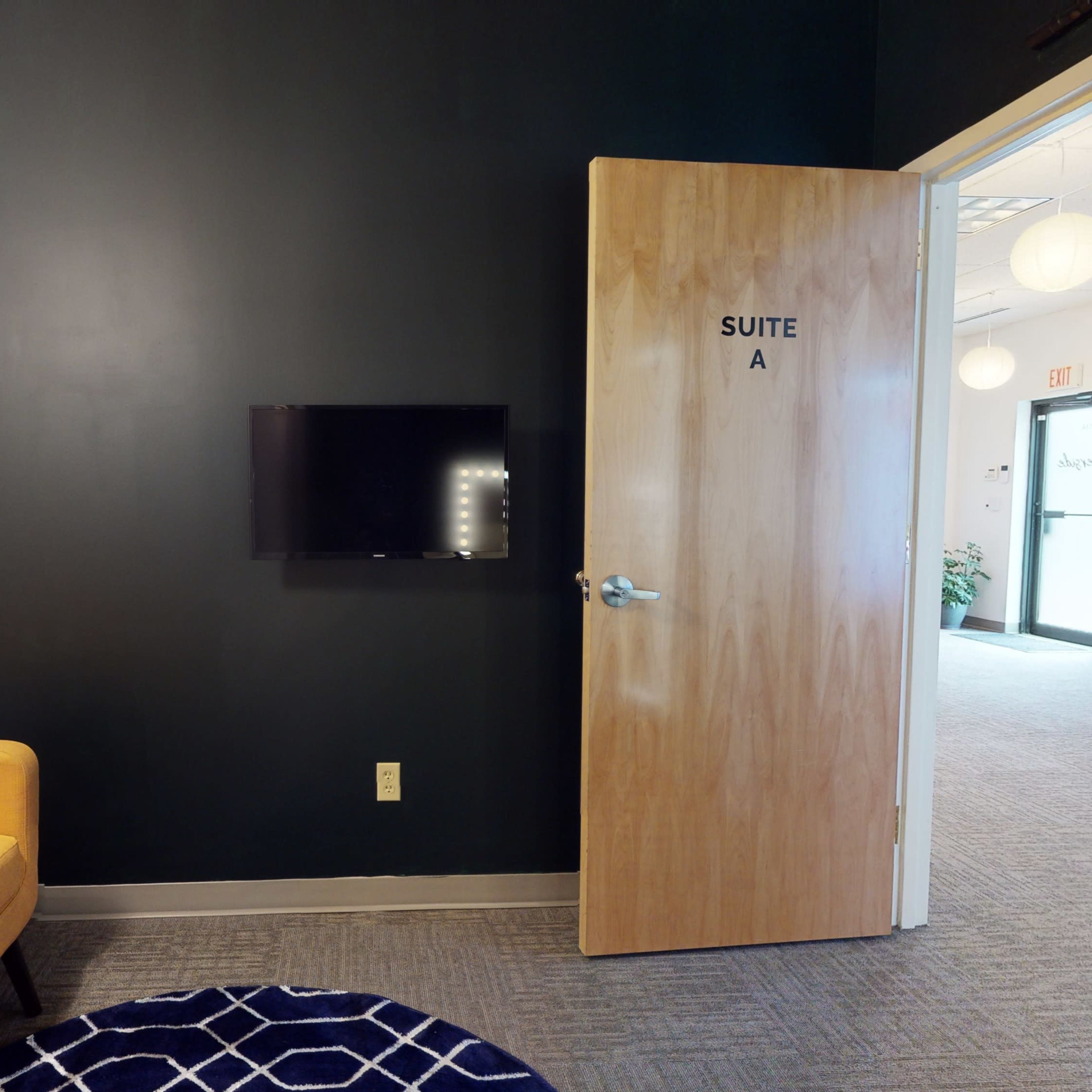 A small waiting room with a yellow sofa, a TV mounted on a black wall, and a door labeled "SUITE A" leading to another area.