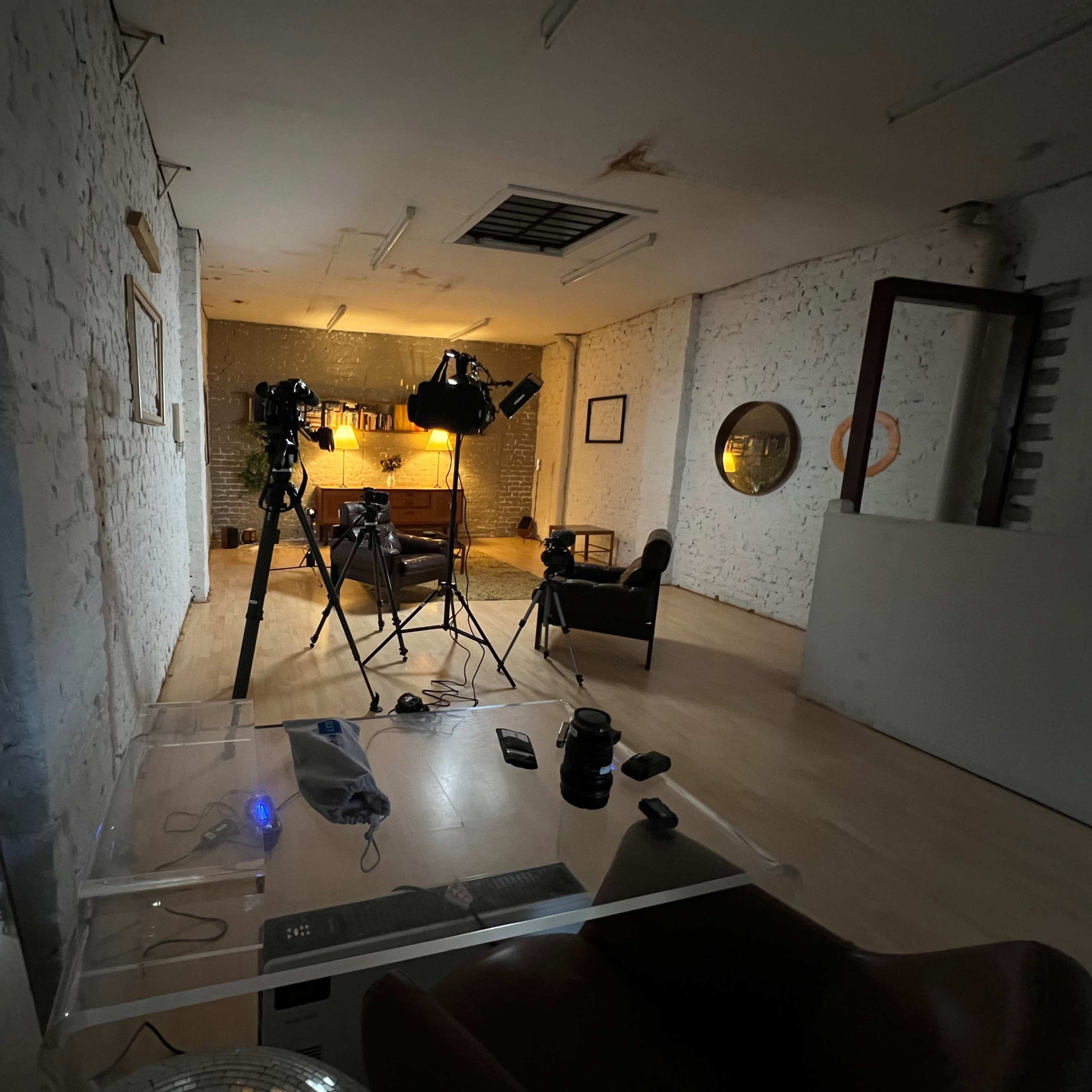 A spacious room with white brick walls, featuring a film setup with cameras, lights, and a few pieces of furniture.