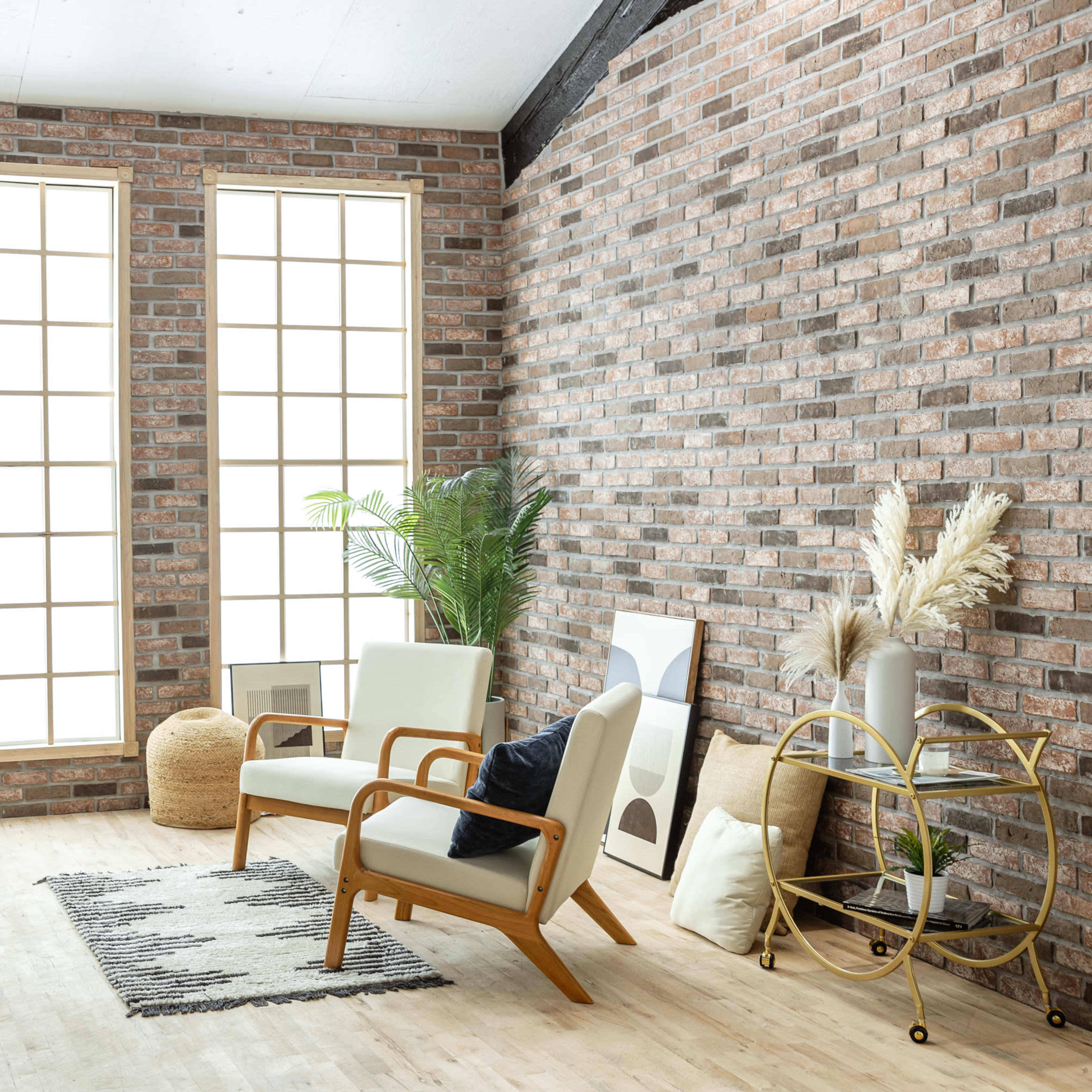 A modern interior space features large windows, a brick wall, a pair of armchairs, a decorative plant, and a gold cart.