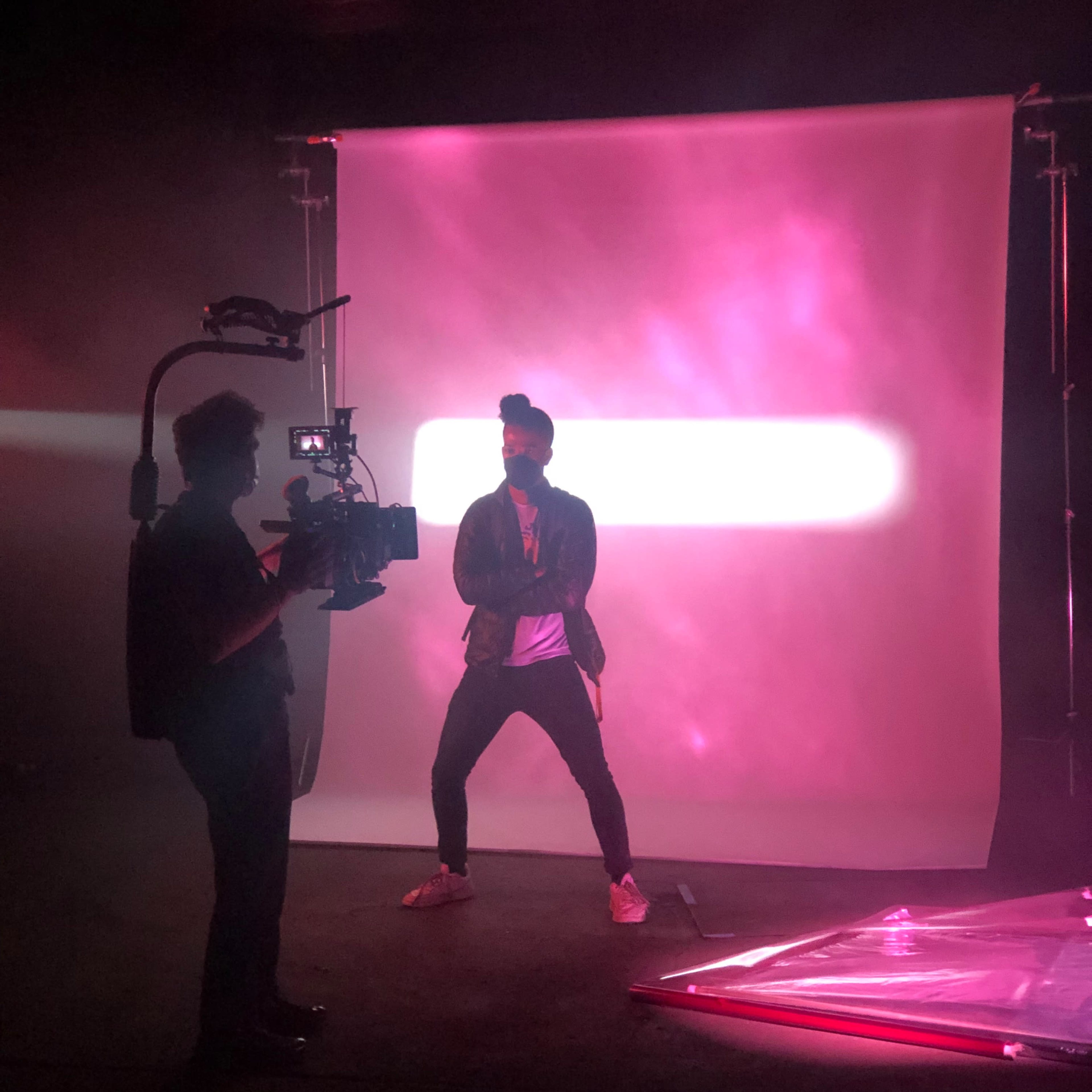 A person poses in front of a backlit pink backdrop while being filmed by another individual holding a camera.
