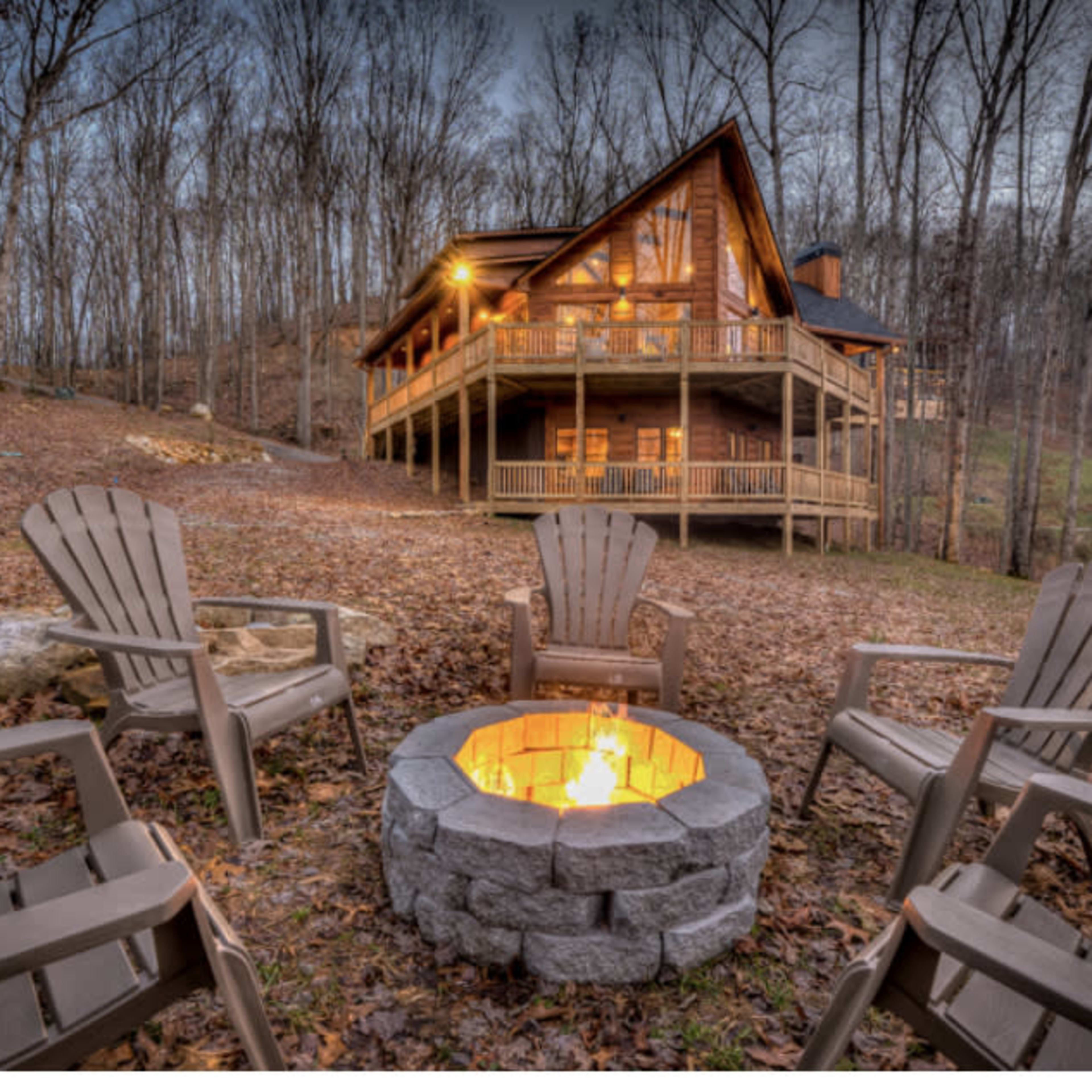 A cozy cabin surrounded by trees features a firepit with four chairs arranged around it.