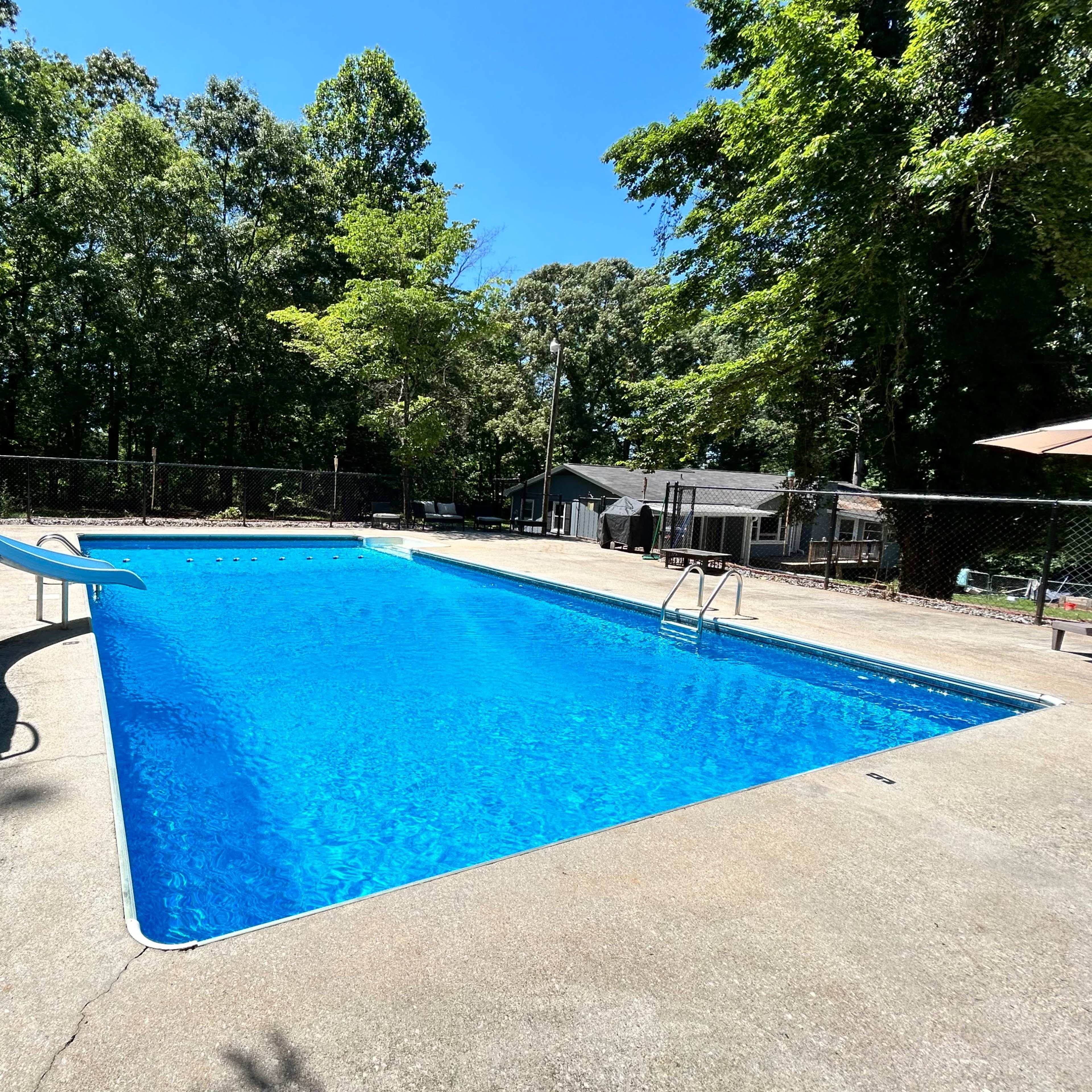 A clear blue swimming pool with a slide and diving board is surrounded by trees and a fence.