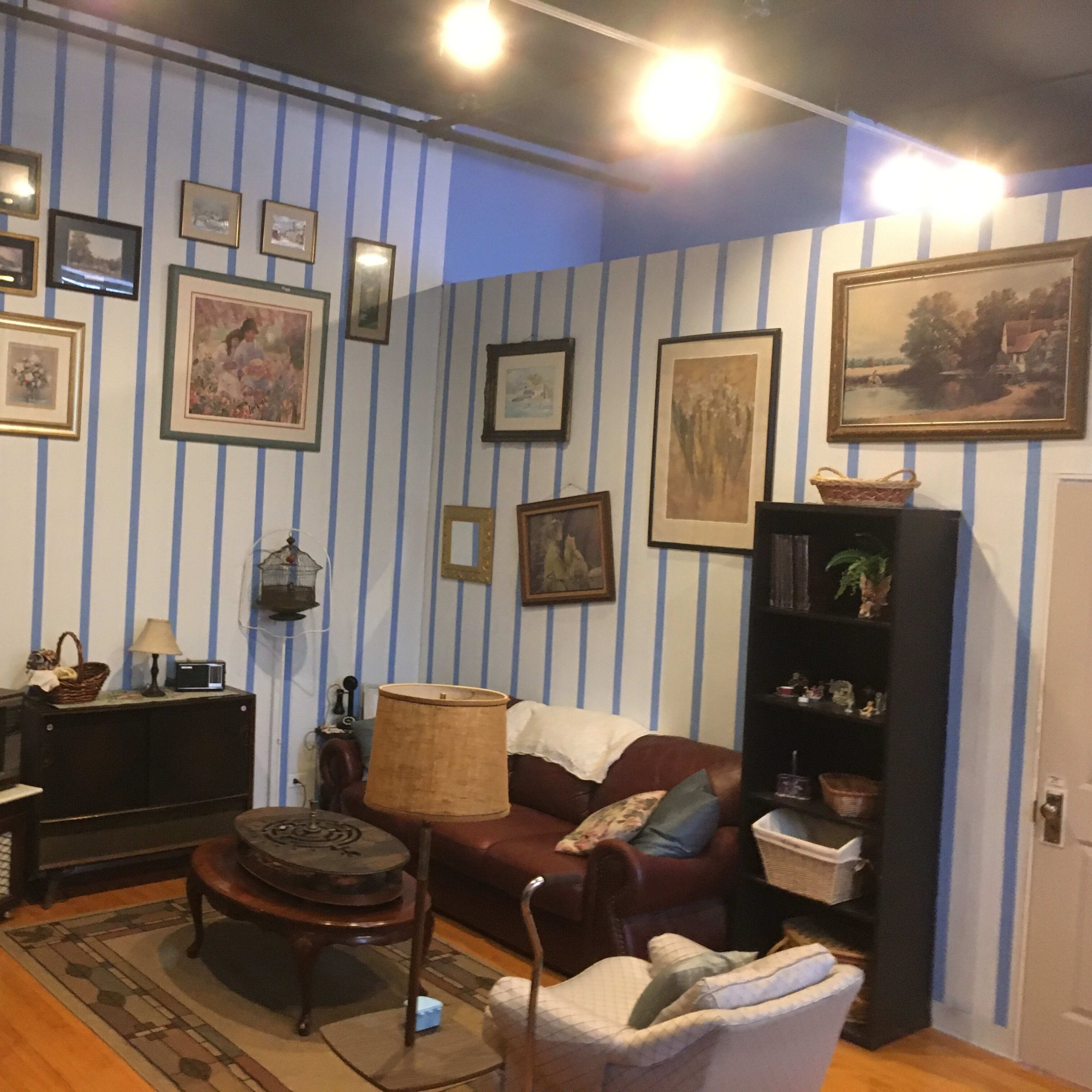 The room features striped walls adorned with various framed pictures, a vintage television set on a wooden cabinet, and a mix of upholstered furniture arranged around a central coffee table.