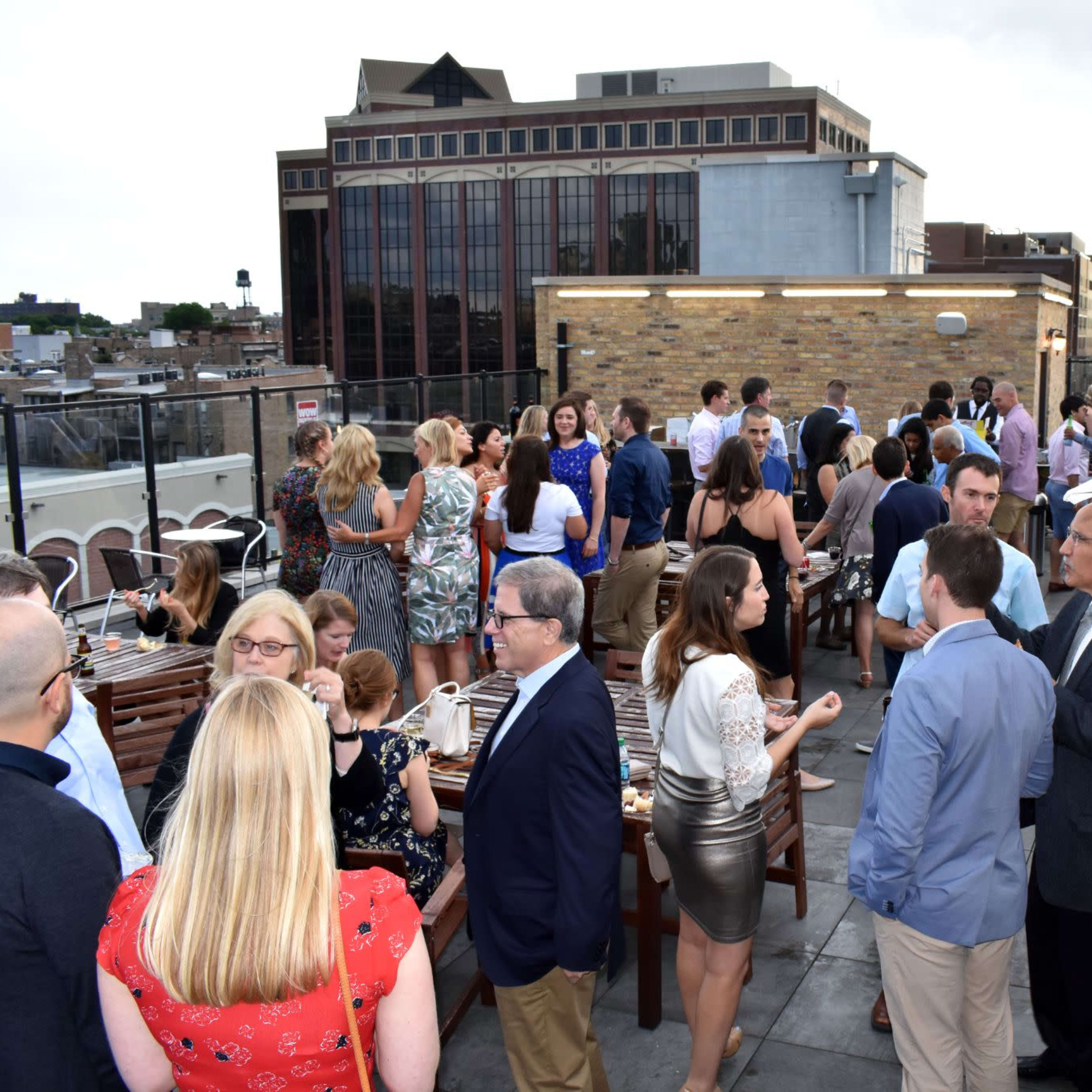 A large group of people mingles on a rooftop, with tables set up for food and drinks against a backdrop of urban buildings.