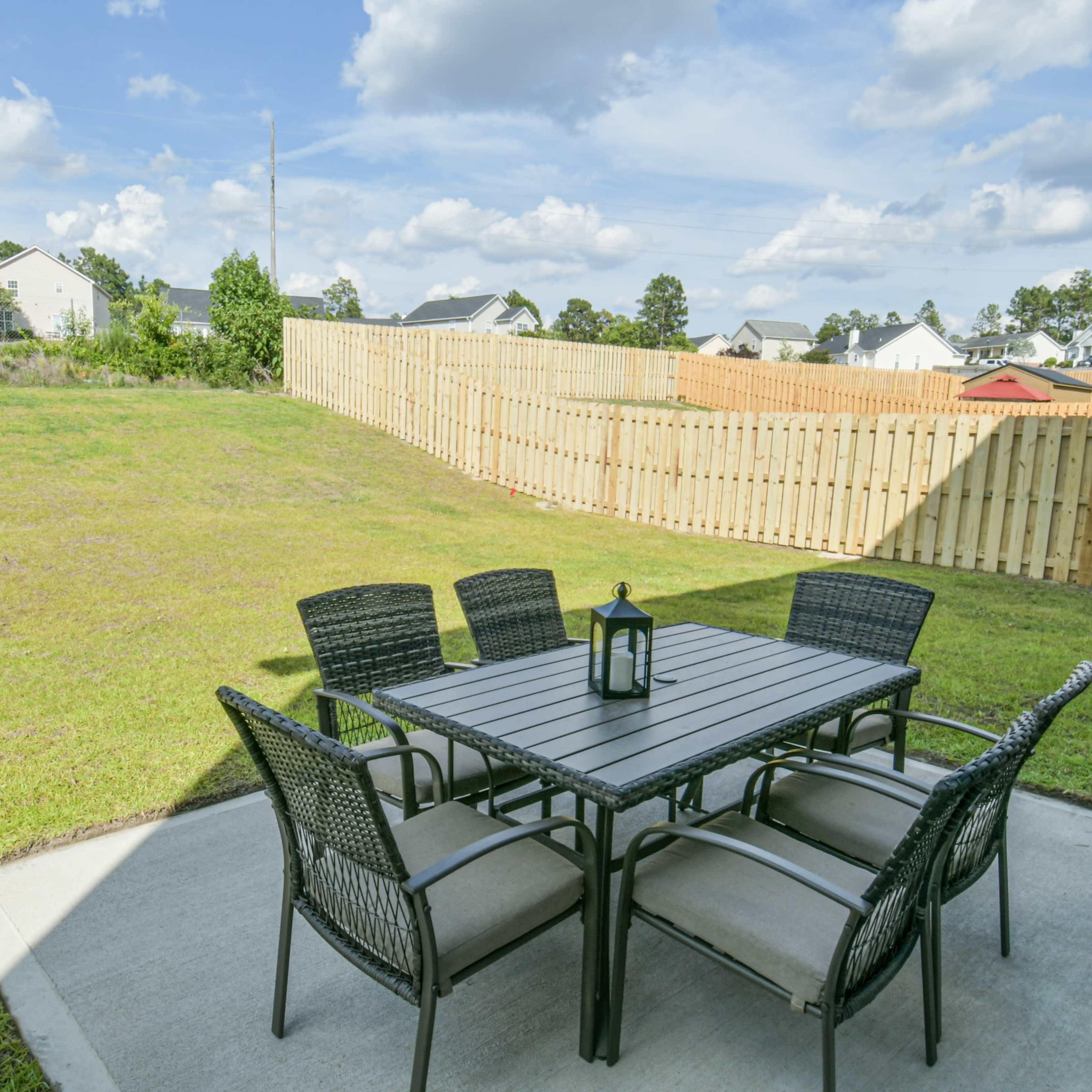 A patio area with a rectangular table and six chairs is set on a concrete slab, surrounded by a grassy lawn and a wooden fence in the background.