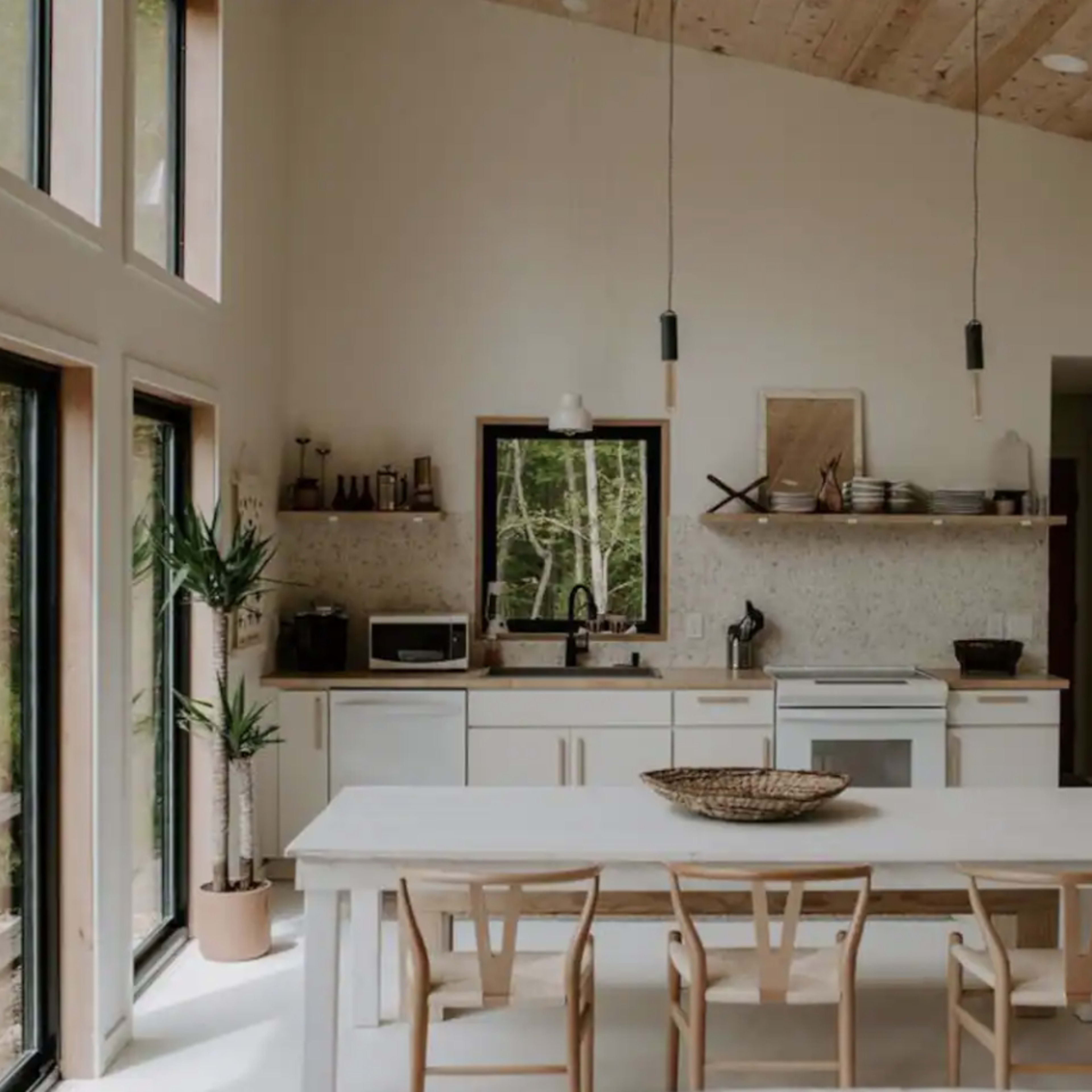 A modern, minimalist kitchen features a large white table surrounded by wooden chairs, with windowed walls allowing natural light and views of greenery outside.