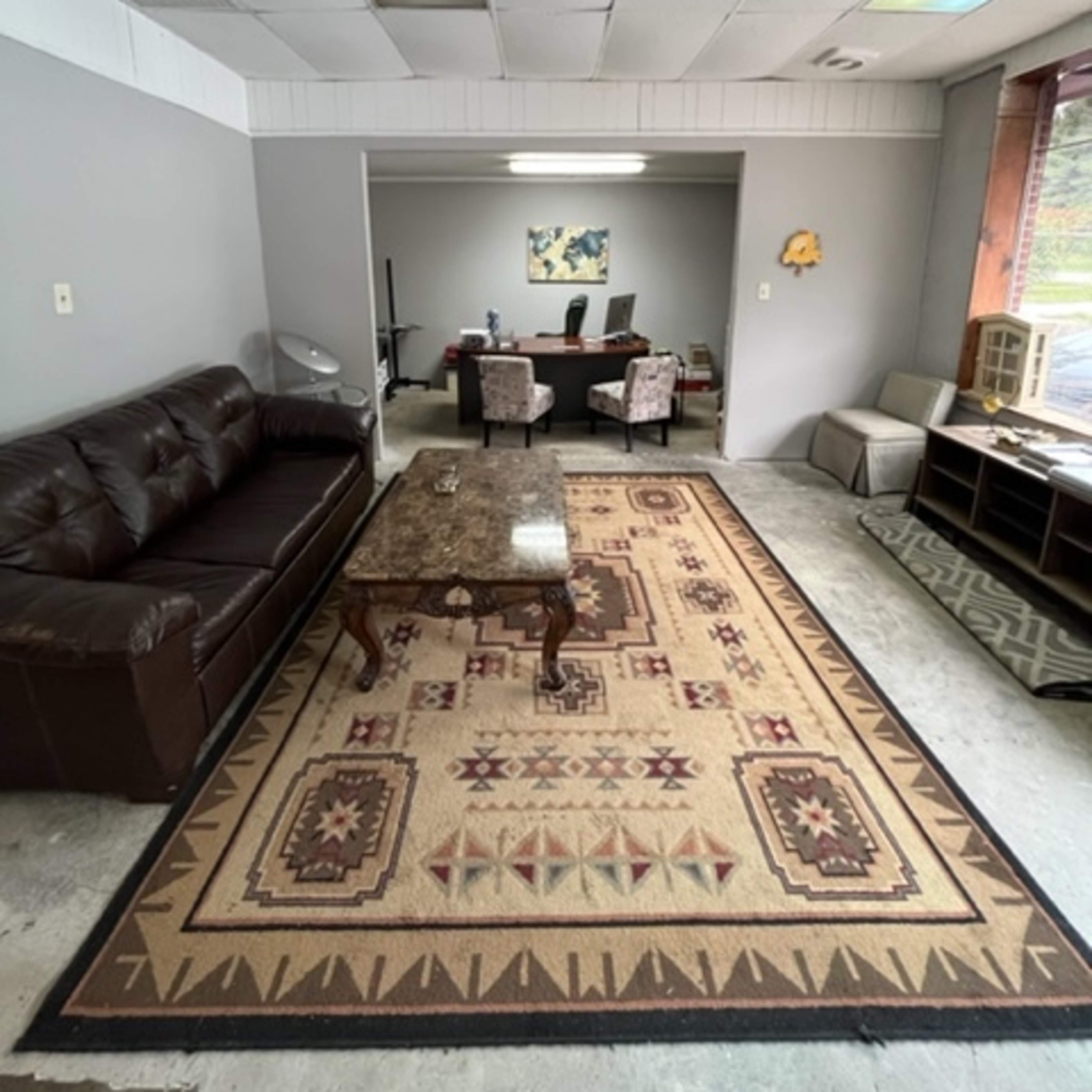 A small office space featuring a brown leather sofa, a patterned area rug, and a view into a separate meeting area with a desk and chairs.