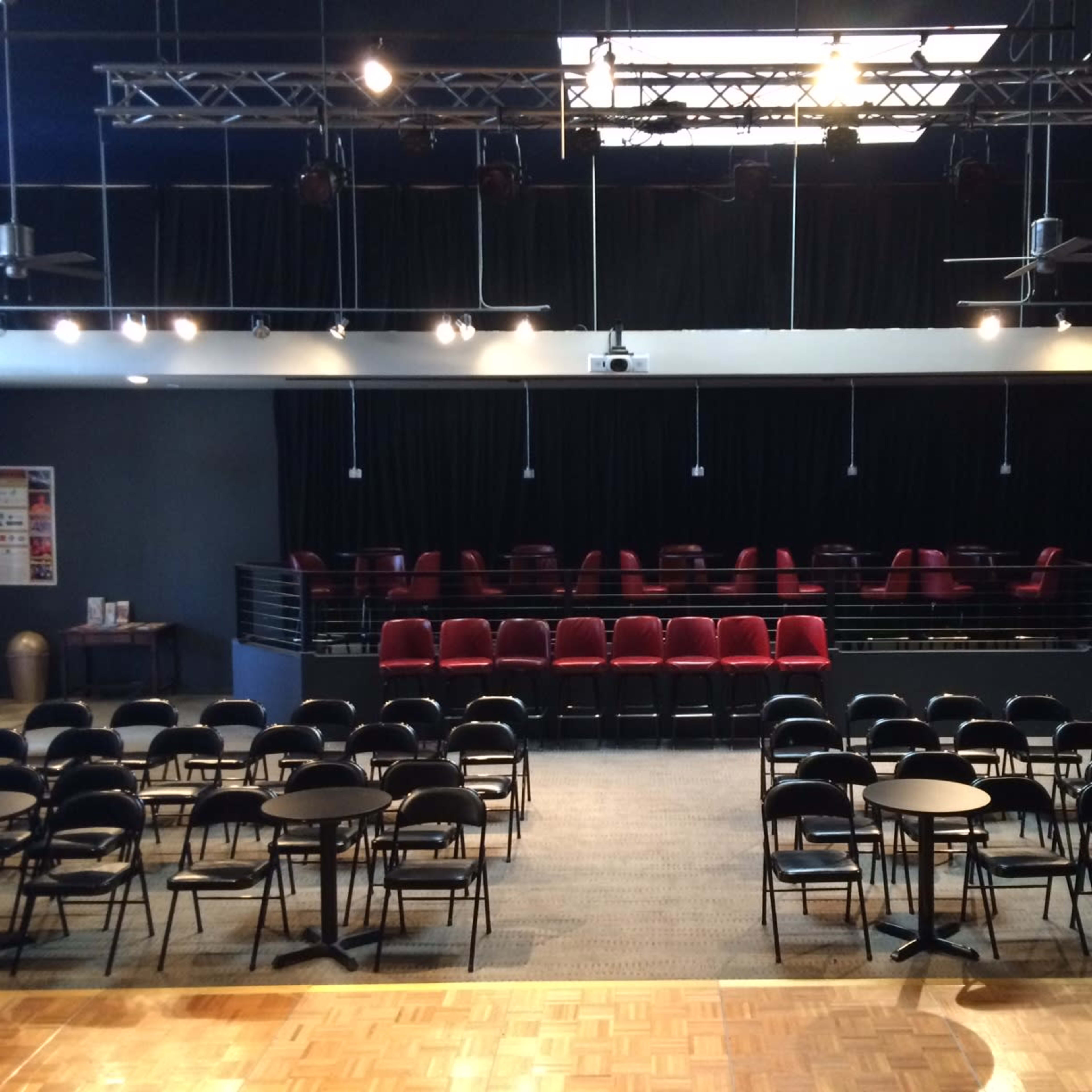 An auditorium with rows of black chairs and round tables set up for a performance, facing a small stage with a backdrop of black curtains.