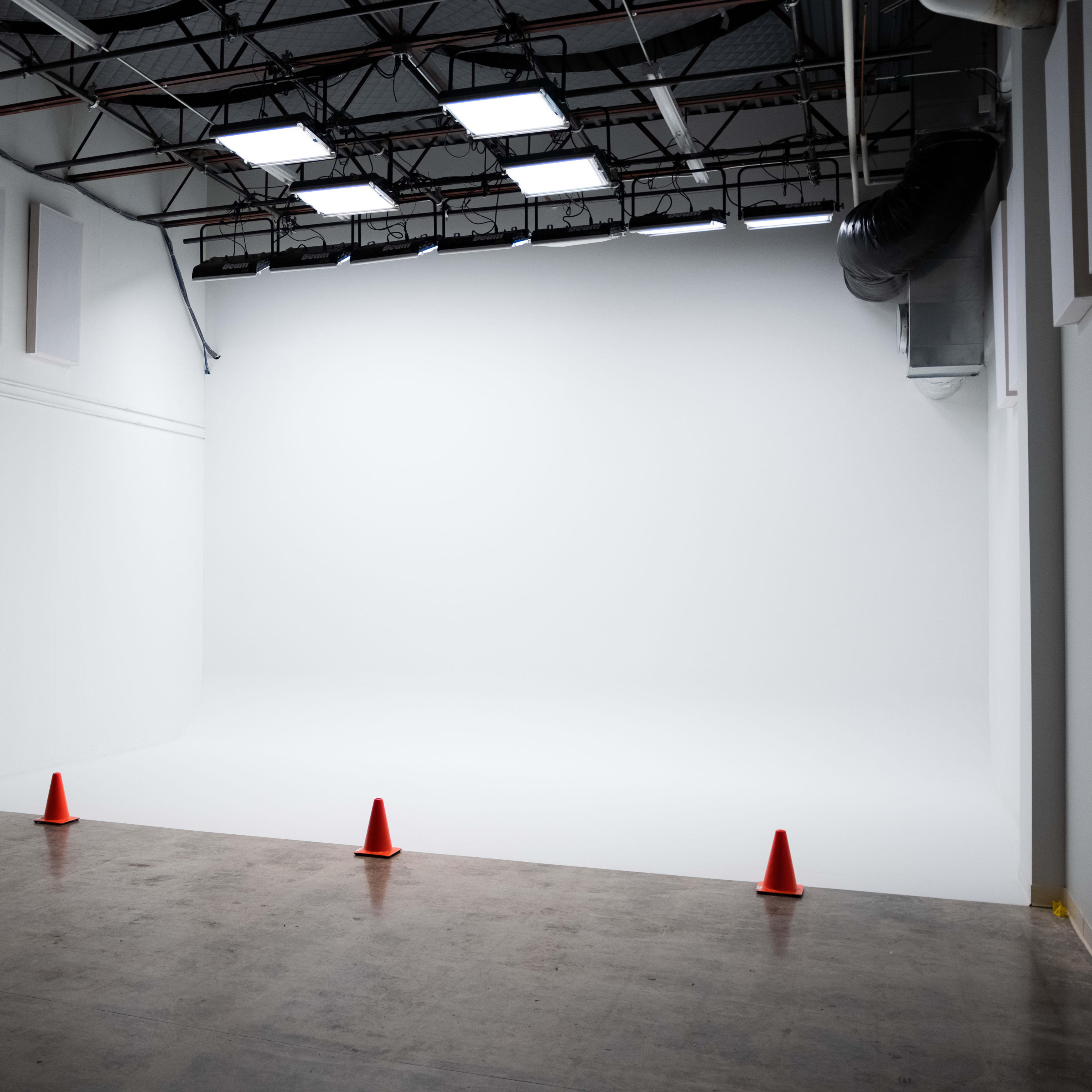 A spacious, empty photography studio with a white backdrop and three traffic cones positioned on the floor.