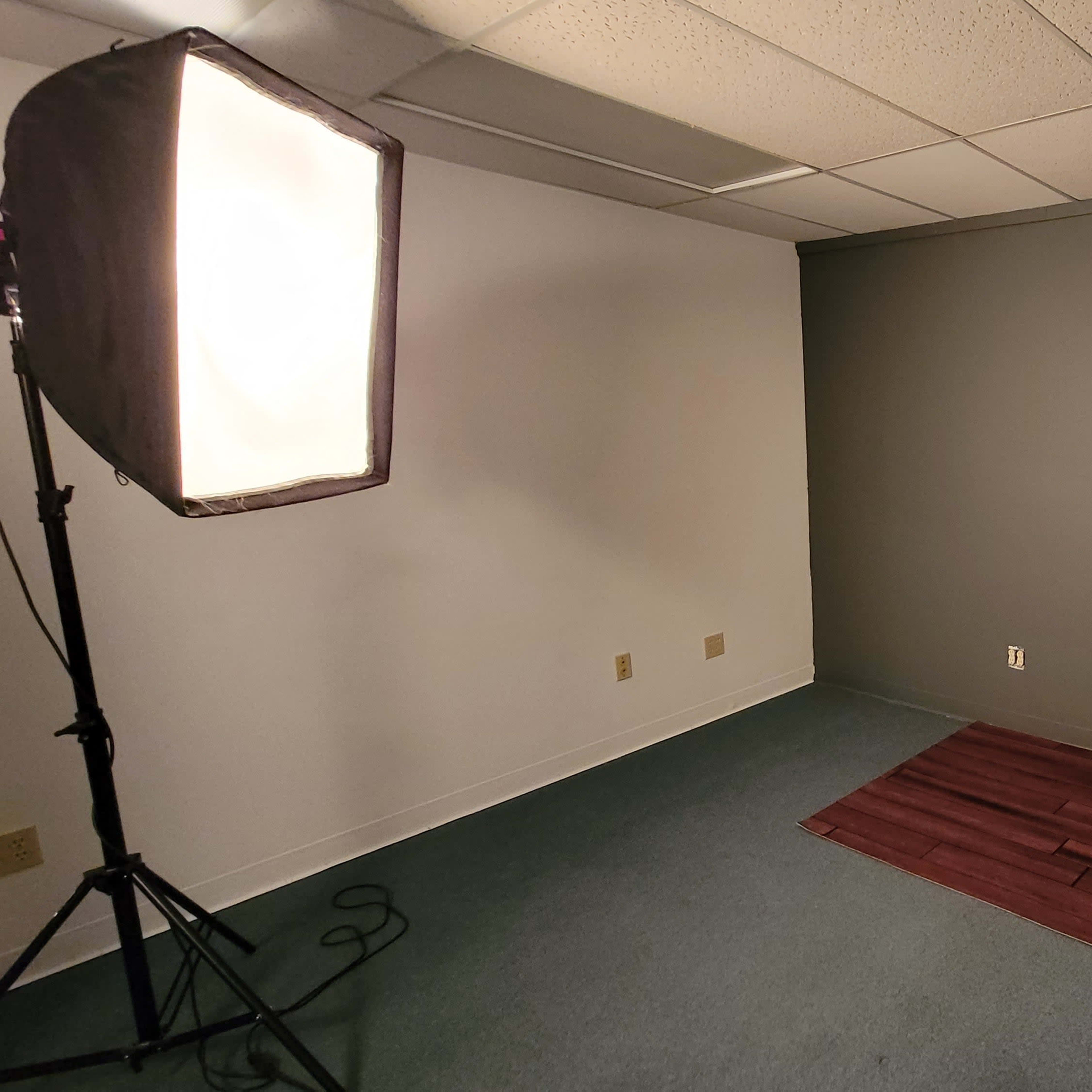 A sparsely furnished studio room with a dark gray wall, a wooden floor section, and two lighting setups on tripods.