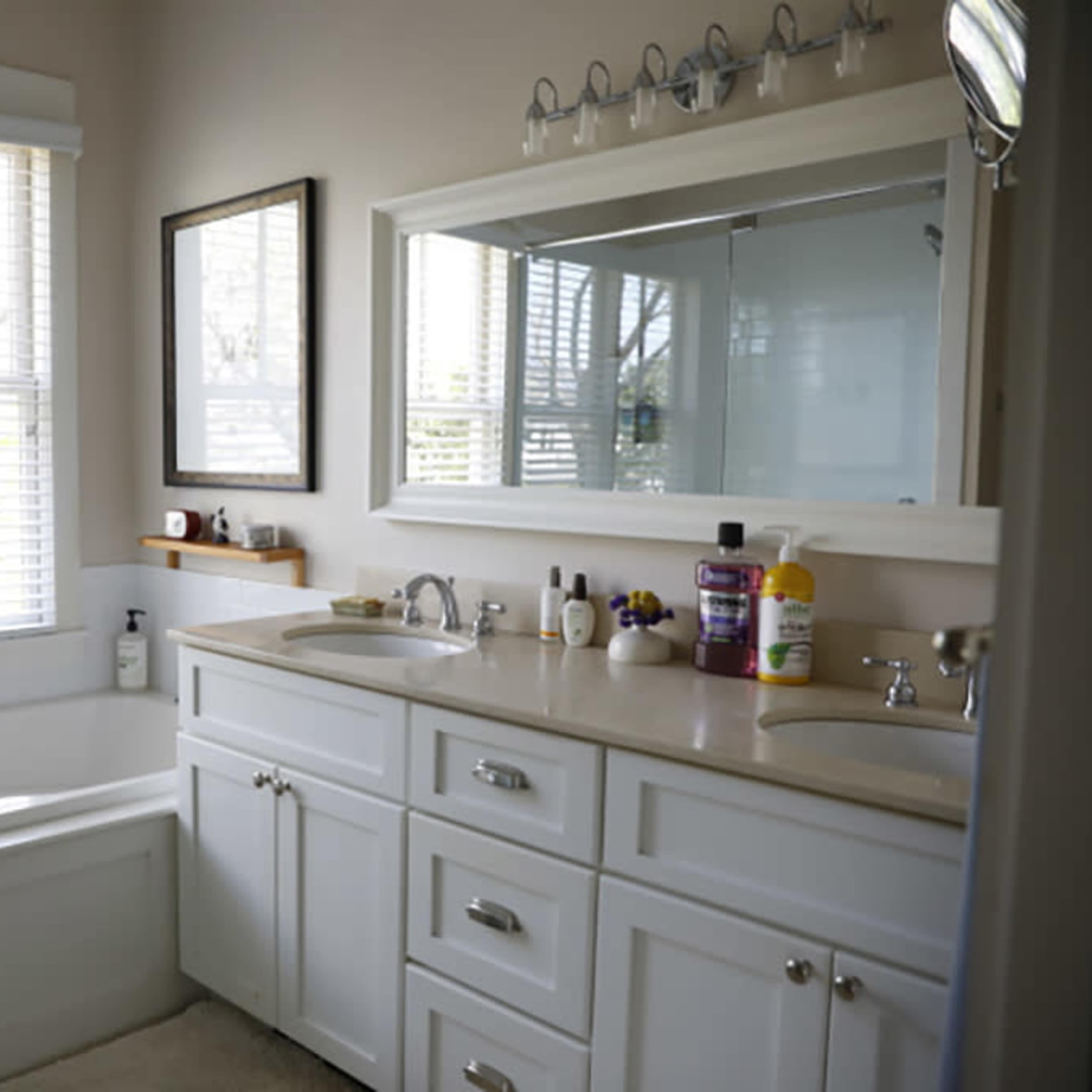 A bathroom featuring a bathtub, a double sink vanity with storage cabinets, a large mirror, and a window with blinds allowing natural light to enter.