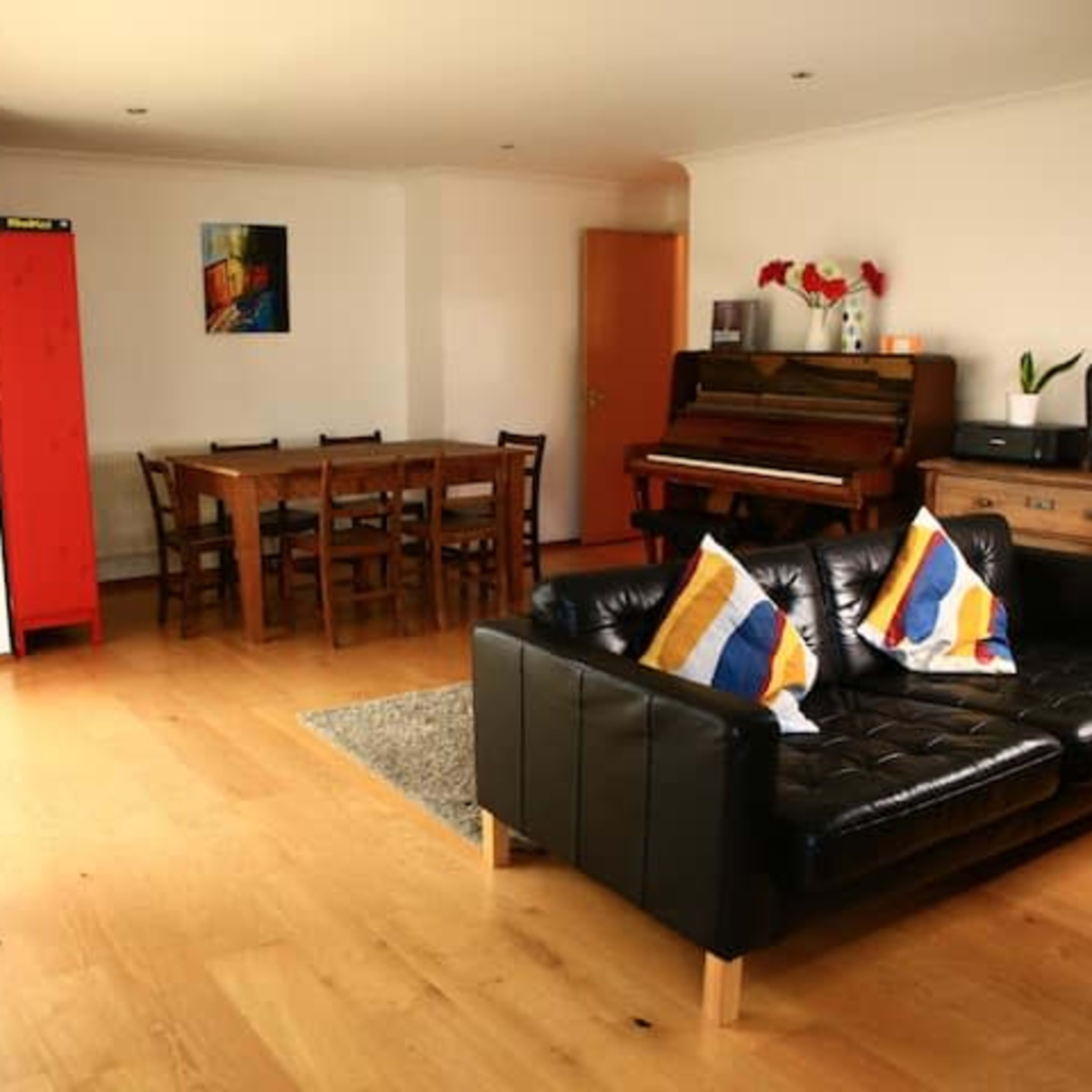 A living area with a black leather sofa, a dining table with chairs, a piano, and a wooden cabinet, all set against a light-colored wall.