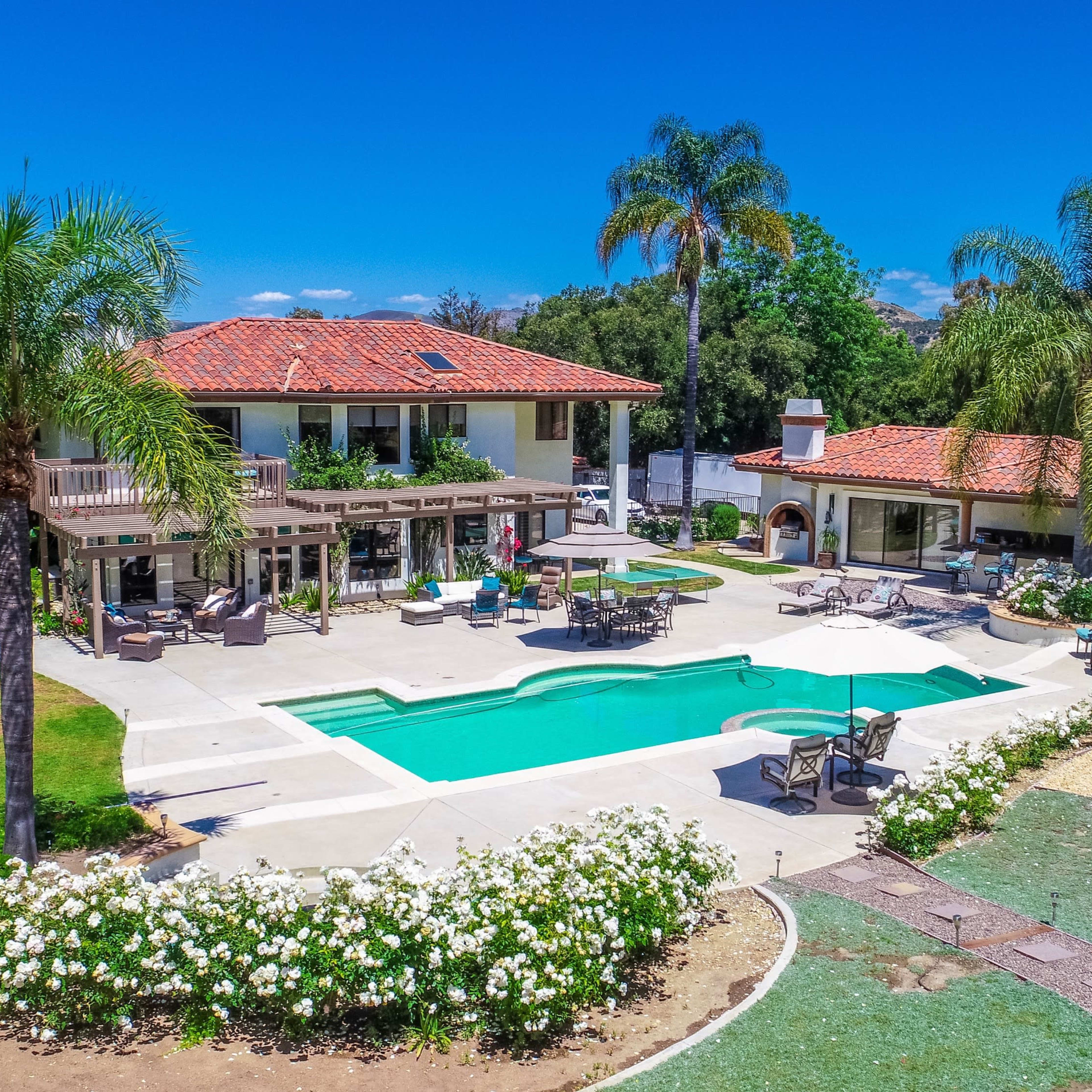 A large property featuring a pool surrounded by a landscaped yard, with a two-story house and an additional structure in the background.
