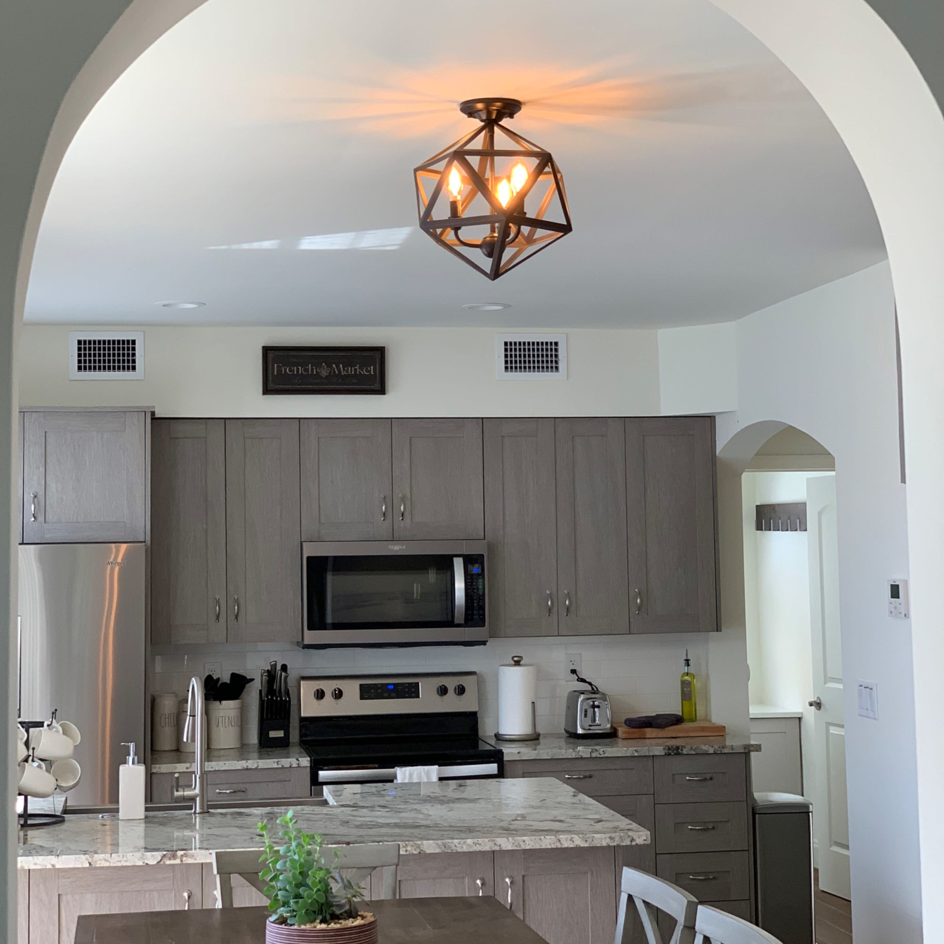 A kitchen features light wood cabinetry, stainless steel appliances, and a geometric pendant light hanging from an arched ceiling.