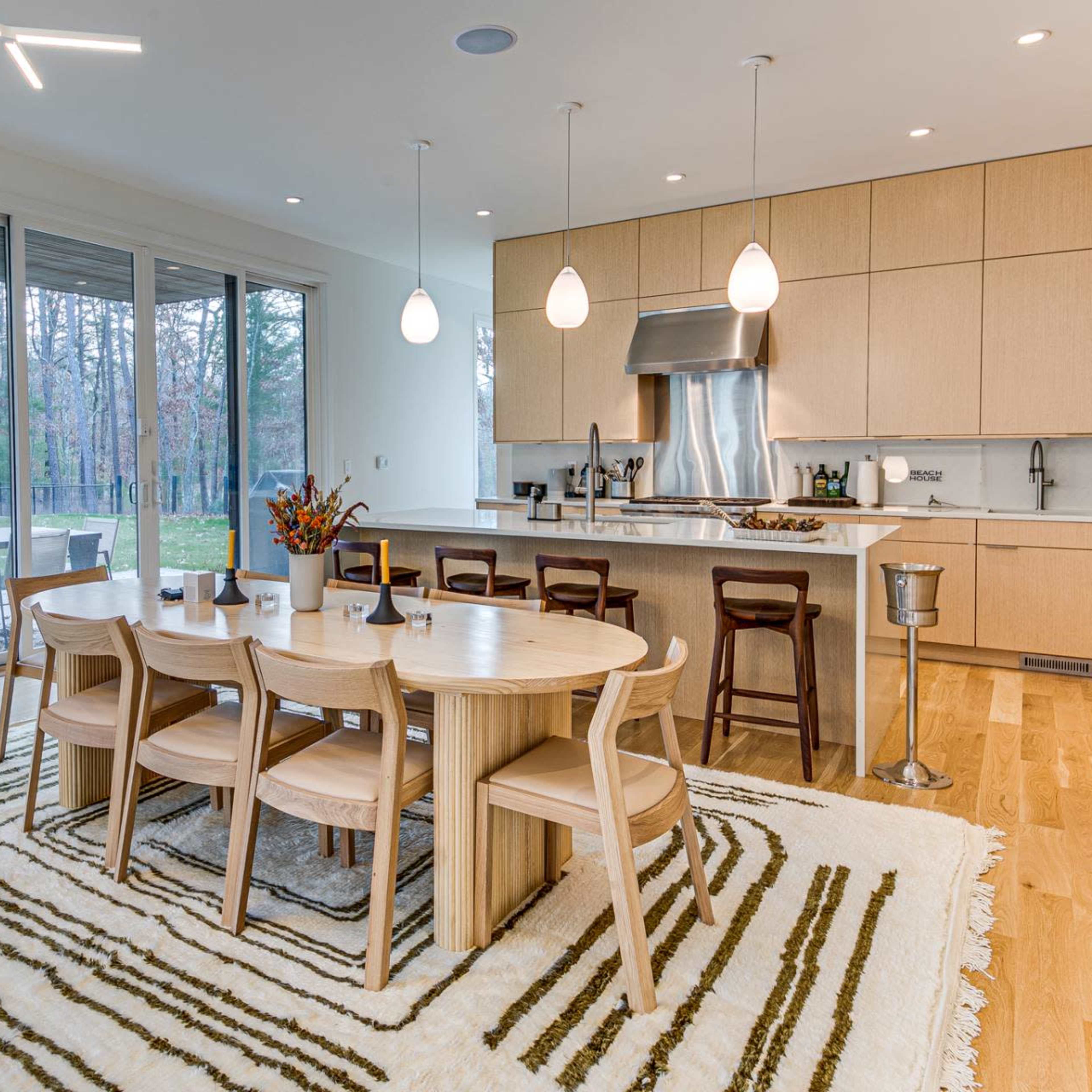A modern kitchen and dining area features a round wooden table with six chairs, surrounded by light-colored cabinetry and large windows that overlook a forested area.