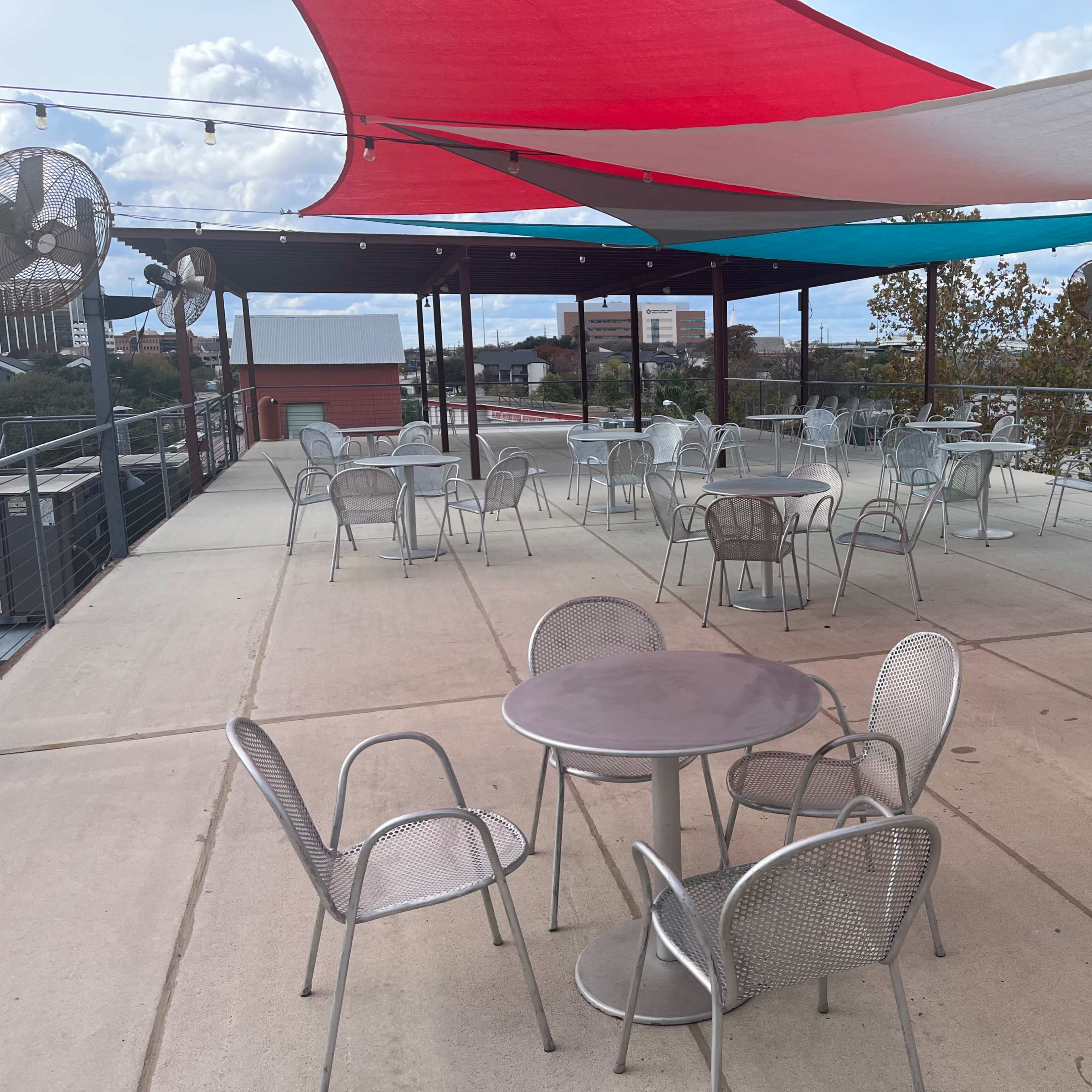 An outdoor dining area with metal tables and chairs under colorful shade sails.