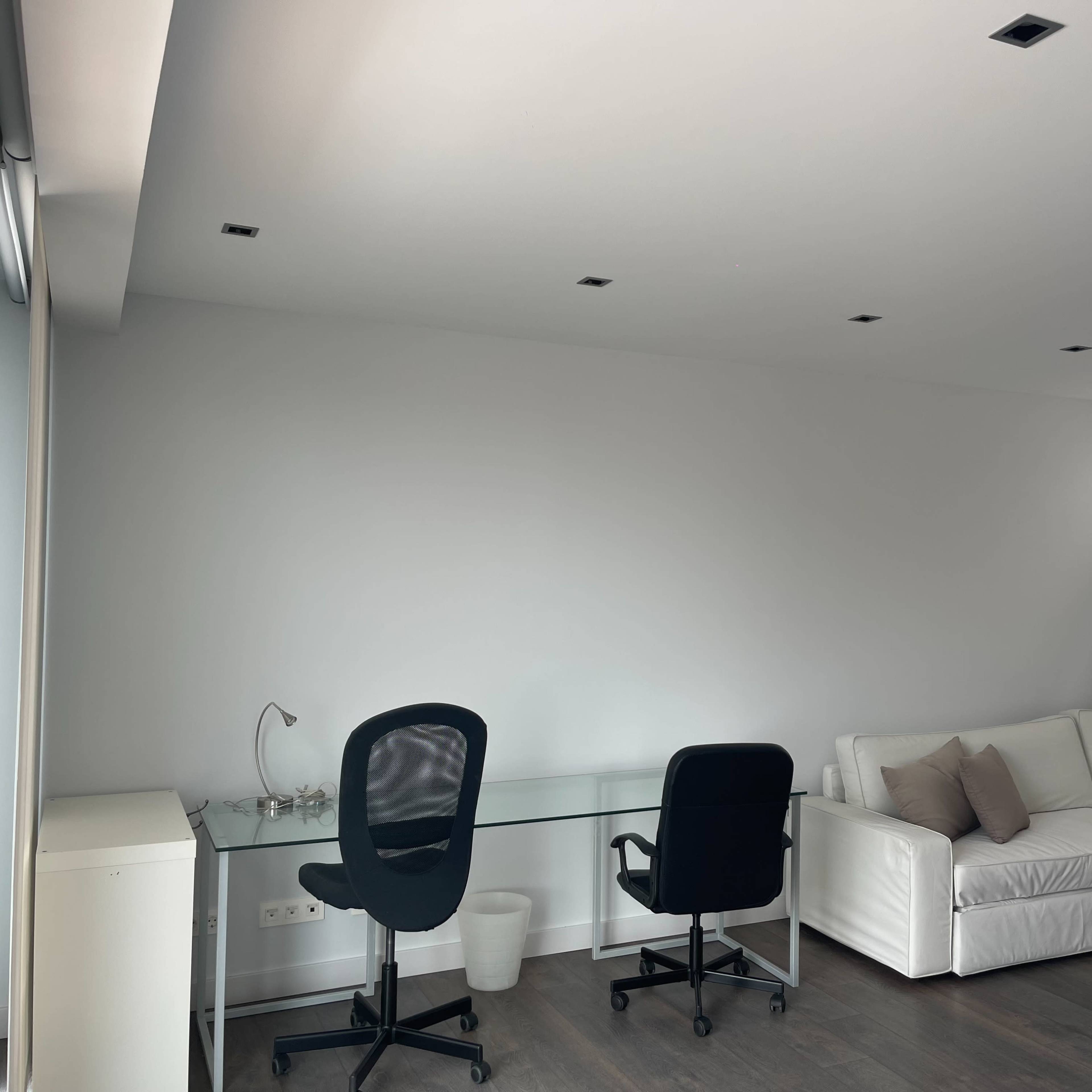 A minimalist room with a glass desk, two black chairs, and a white sofa against a light gray wall.