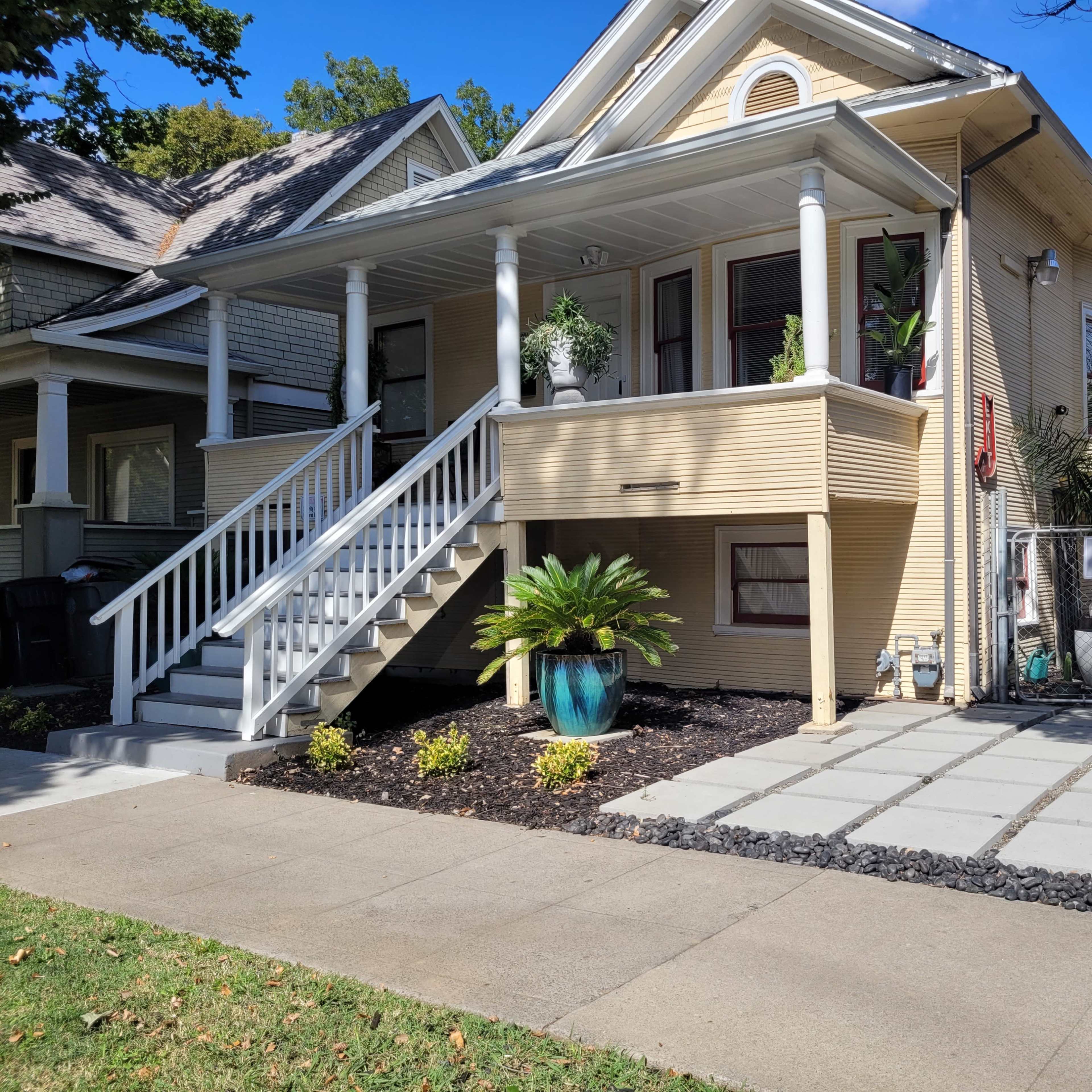 A two-story house with a front porch, a white staircase, and a landscaped yard featuring potted plants and a stone pathway.
