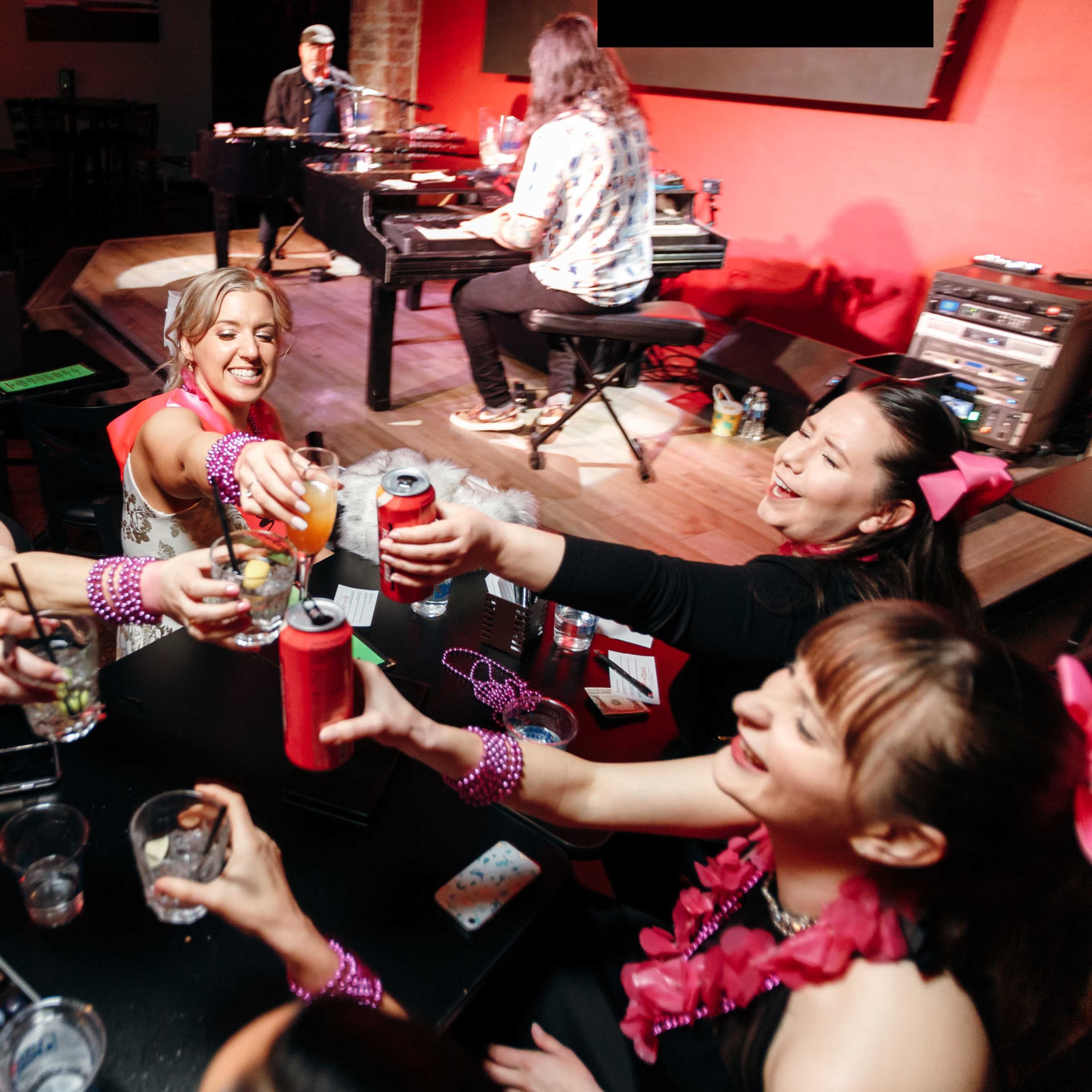 A group of women in festive attire raise their drinks in a lively bar while a musician plays a piano on stage.