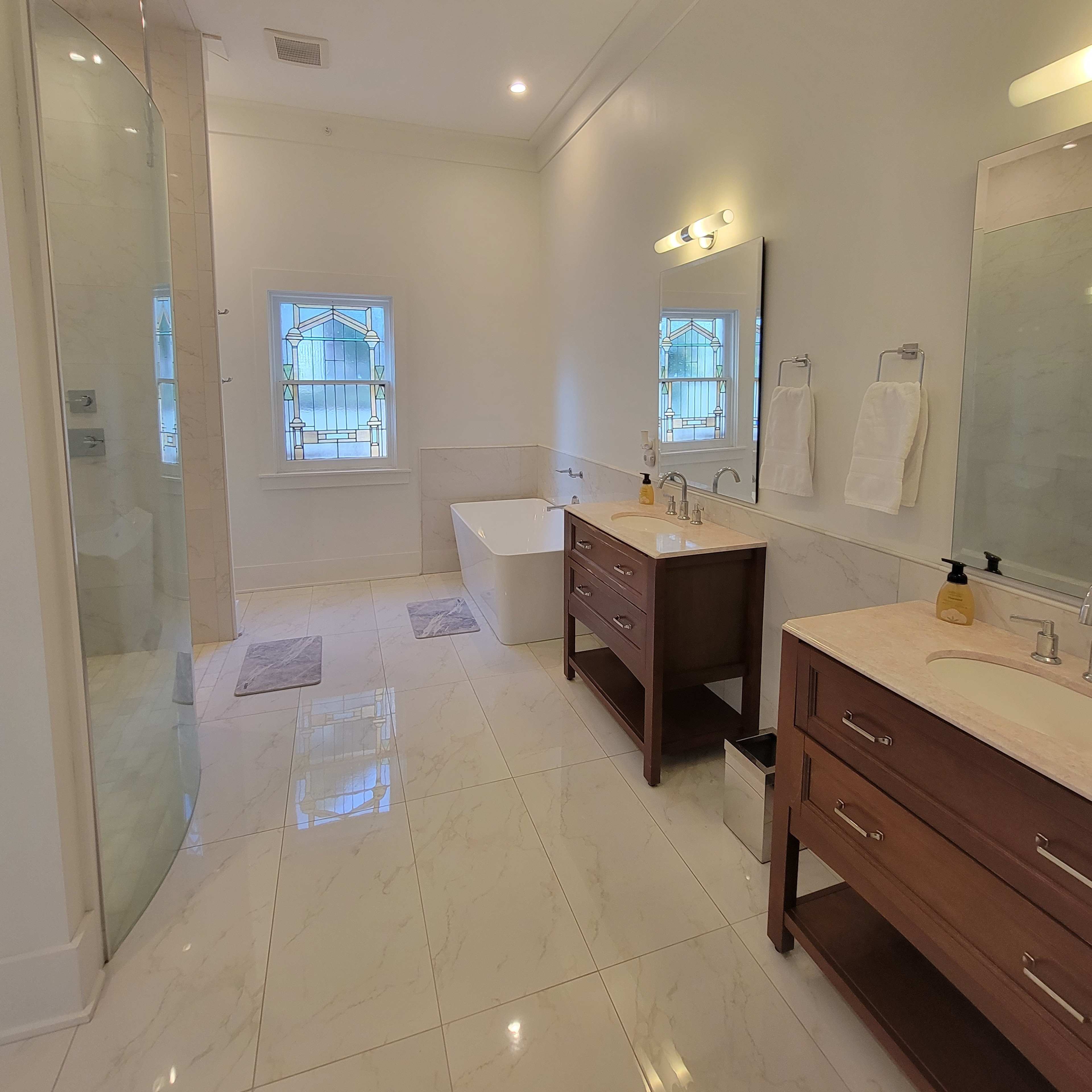 A modern bathroom featuring a glass shower enclosure, a freestanding bathtub, two sinks with wooden vanities, and large windows.