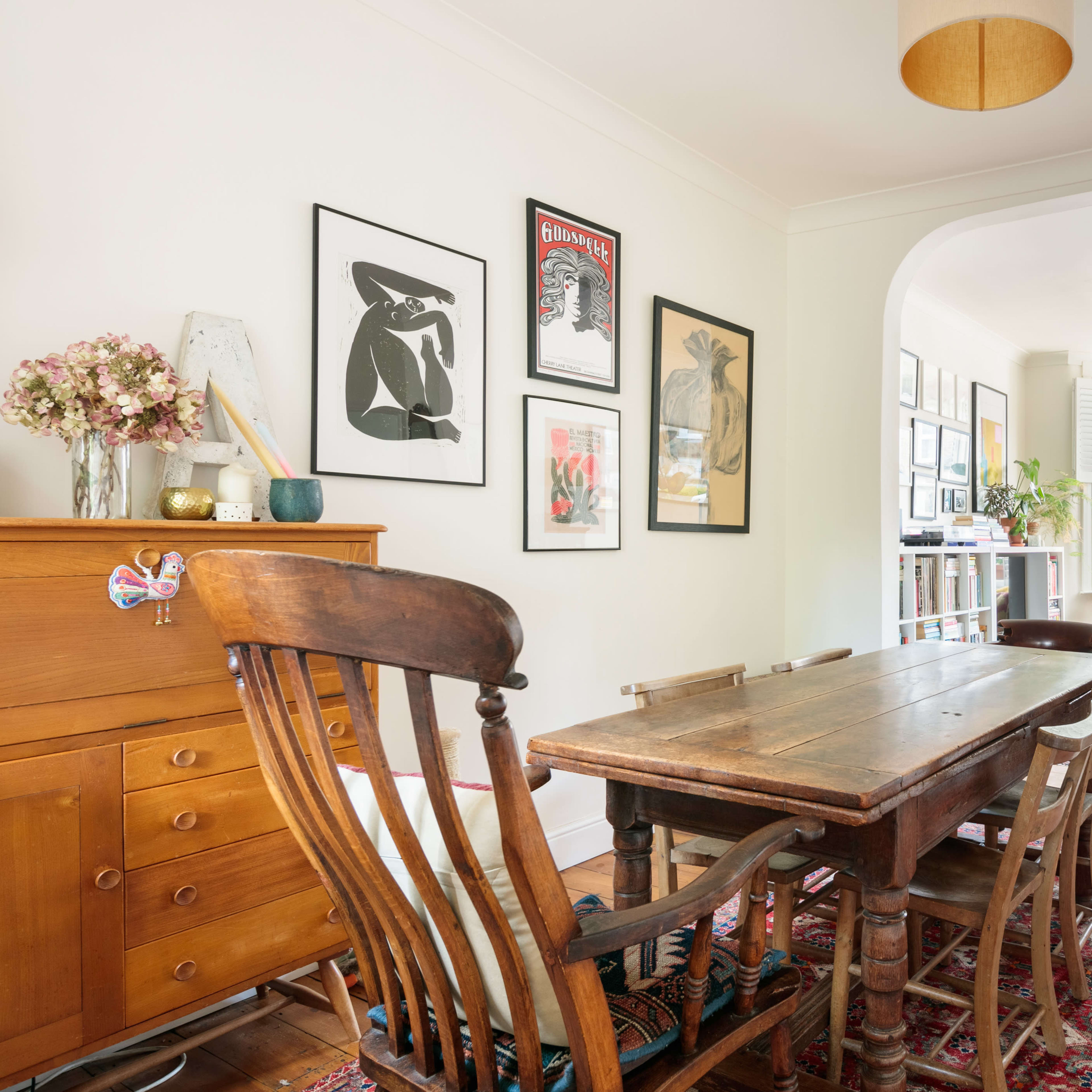 A bright dining area with a wooden table surrounded by chairs, framed artwork on the walls, and a view into a cozy, well-lit living space.