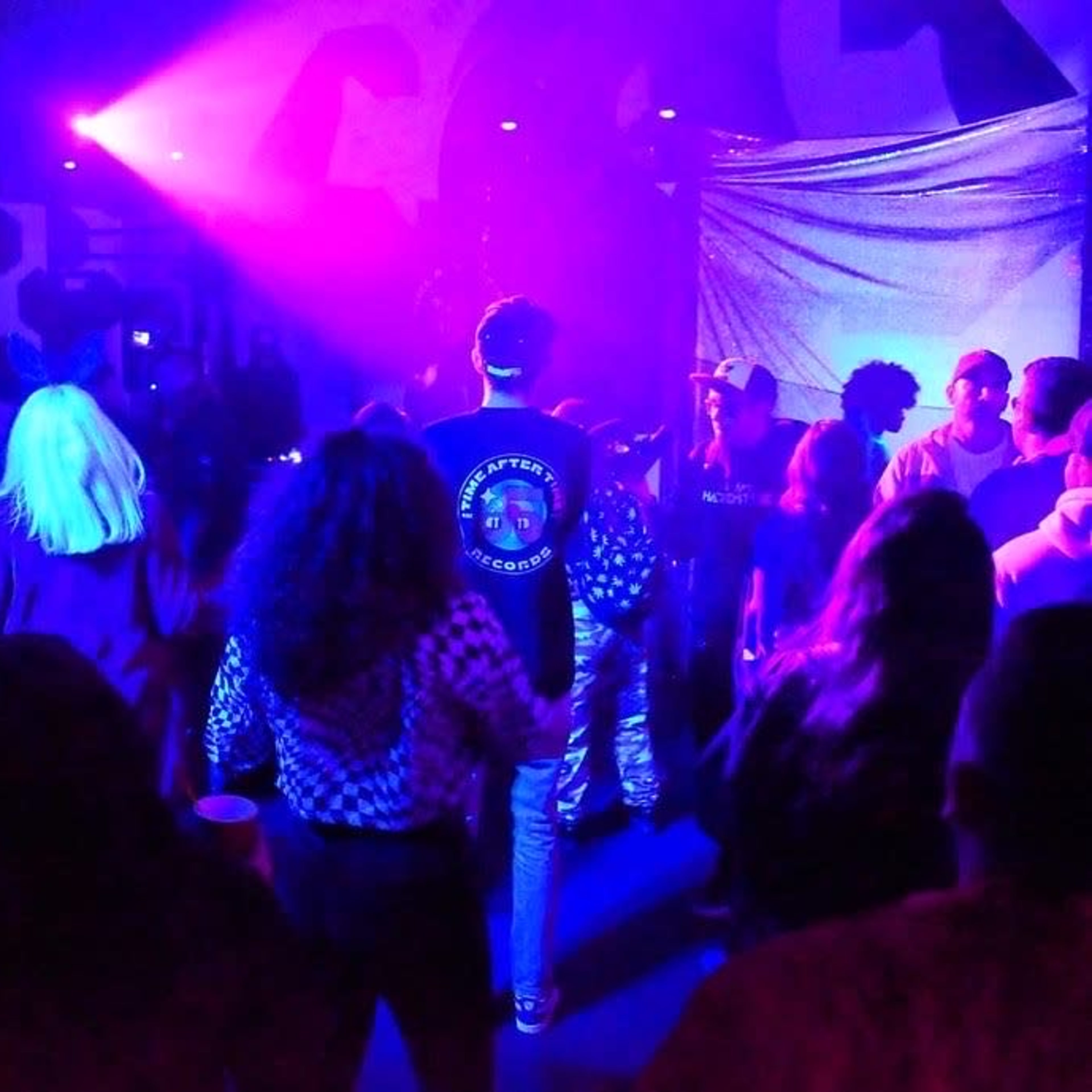 A crowd of people dances under purple and blue lights in a lively indoor setting.