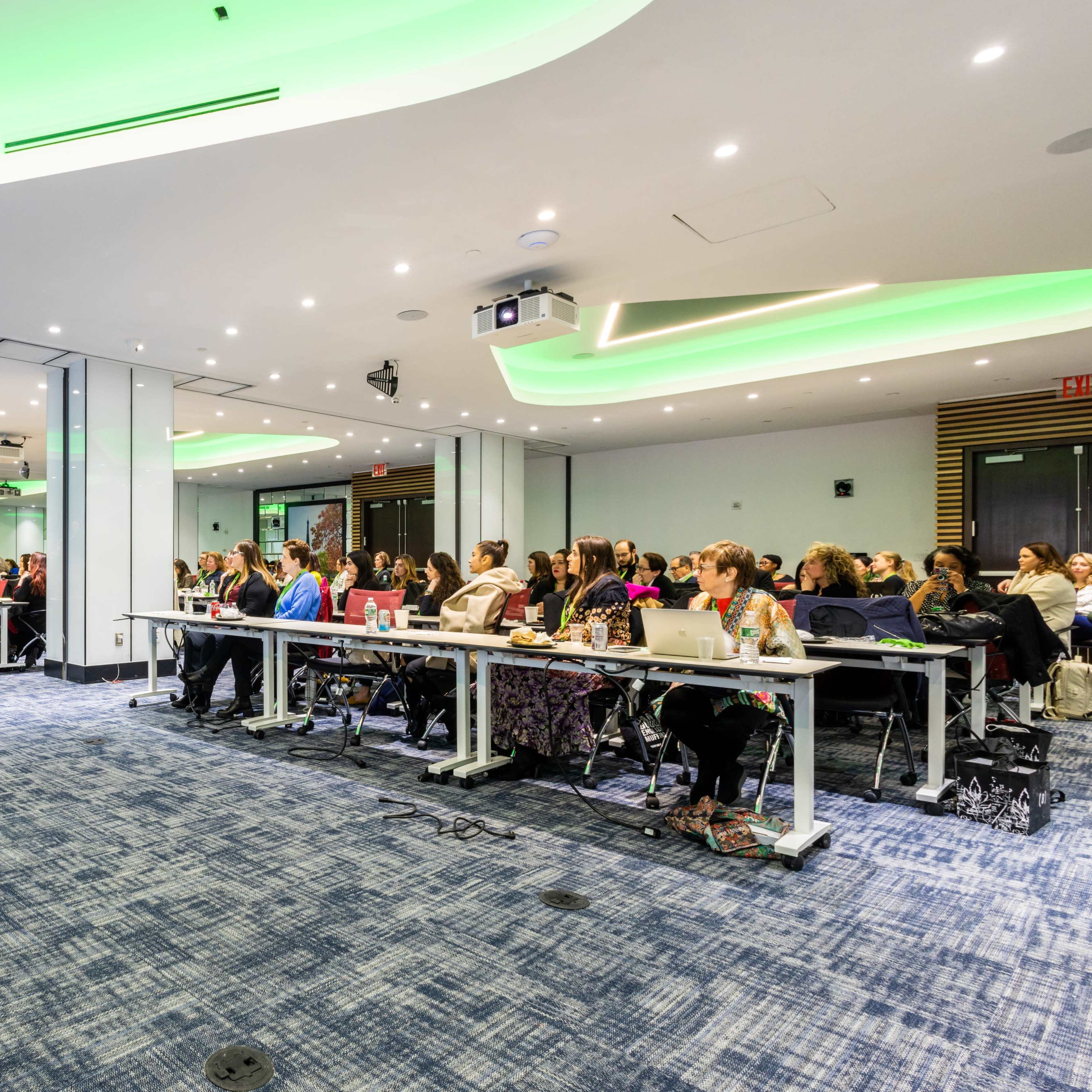 A group of people sits at rows of tables in a conference room, facing a presenter on stage under green-lit ceilings.