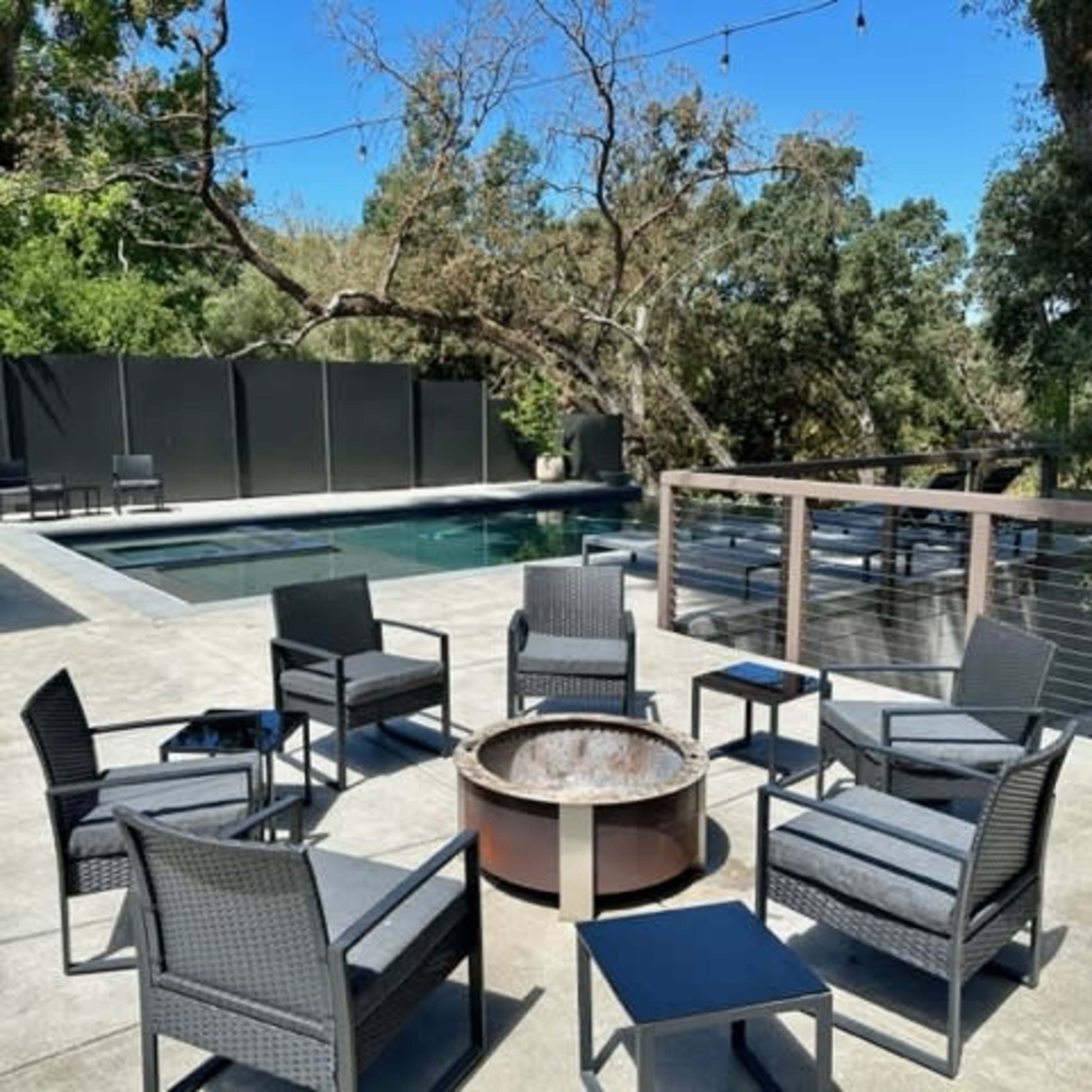 A seating area with six chairs arranged around a fire pit overlooks a pool surrounded by trees and a deck.