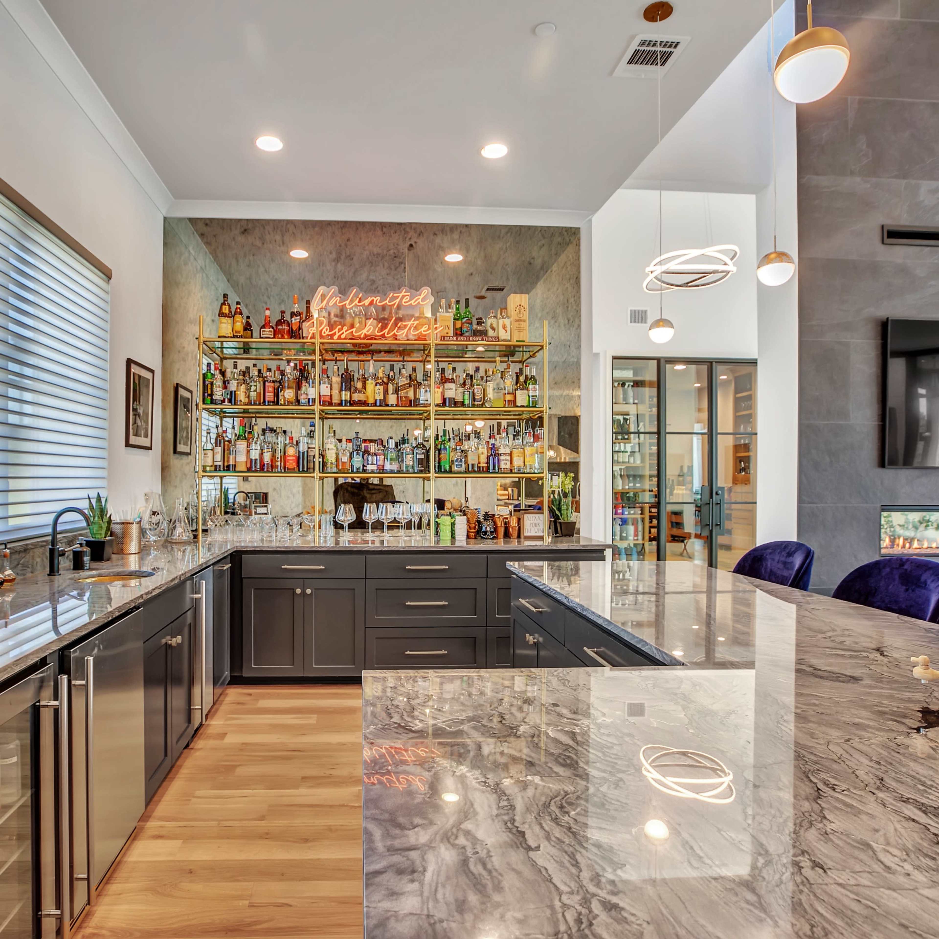 A modern kitchen features a marble countertop, dark cabinetry, and a well-stocked bar with a neon sign in the background.