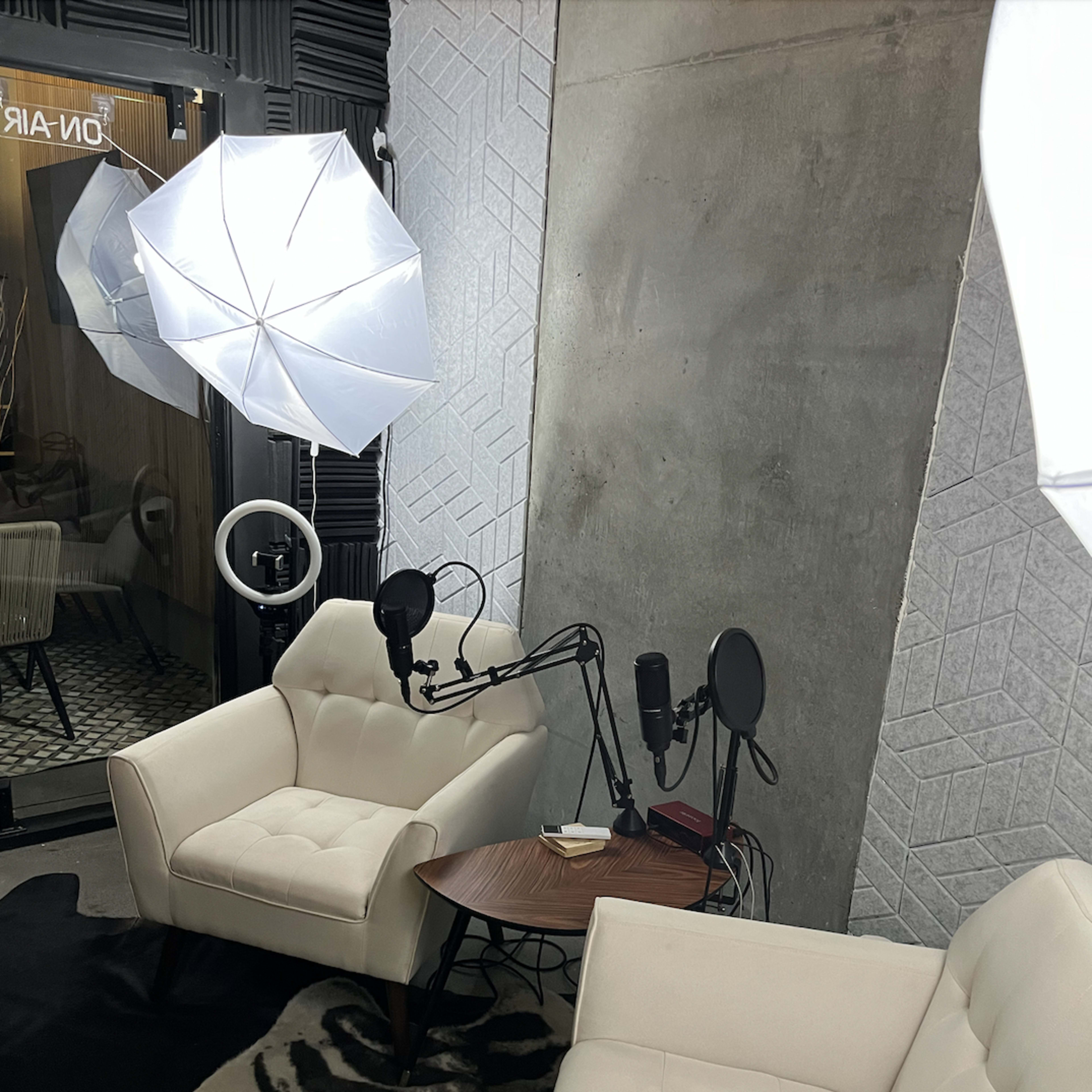 A podcast recording setup with two white chairs, a small round table, and two microphones, surrounded by soft lighting equipment.