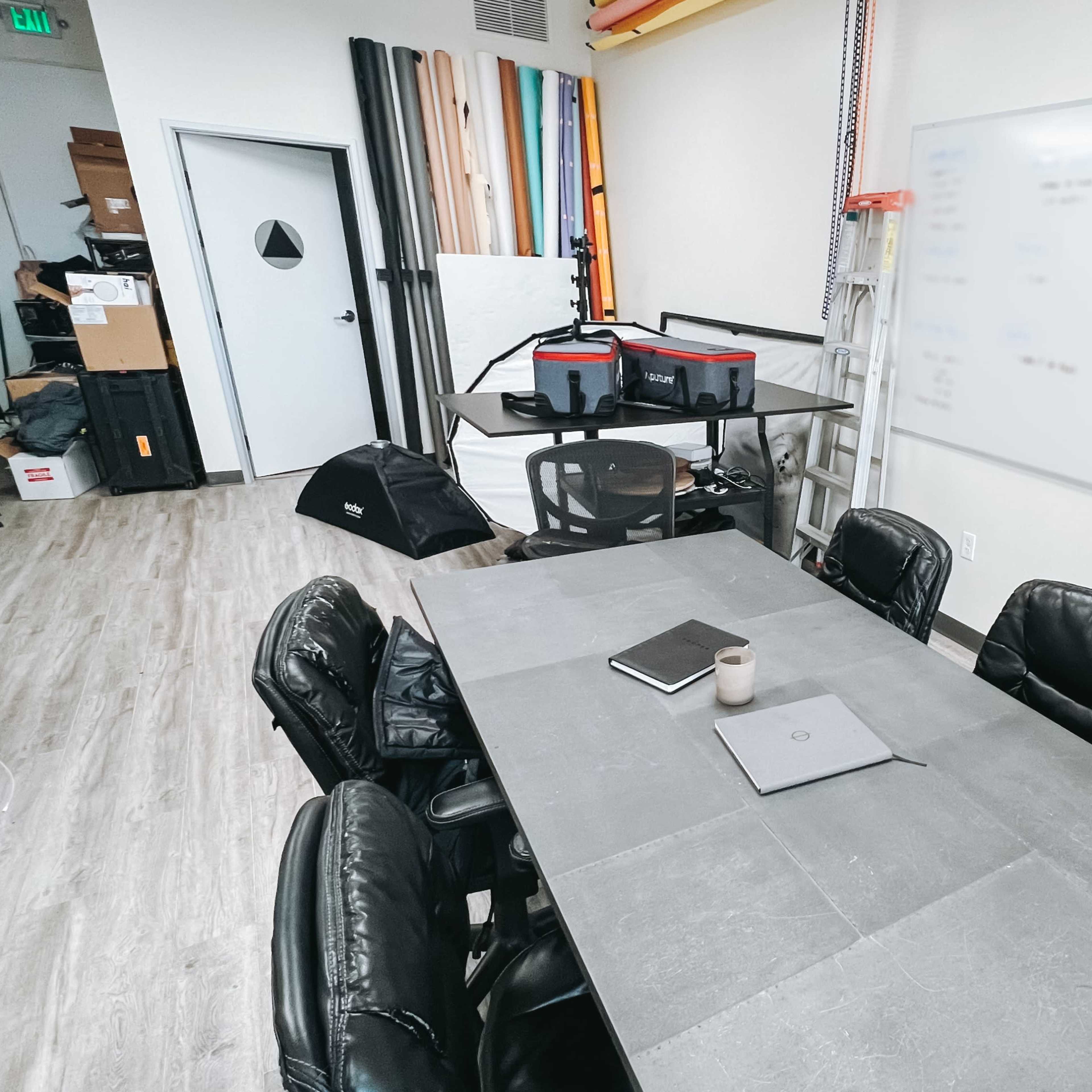A modern conference room features a rectangular table with six black leather chairs, a laptop, and a whiteboard with notes, surrounded by storage and equipment in the background.