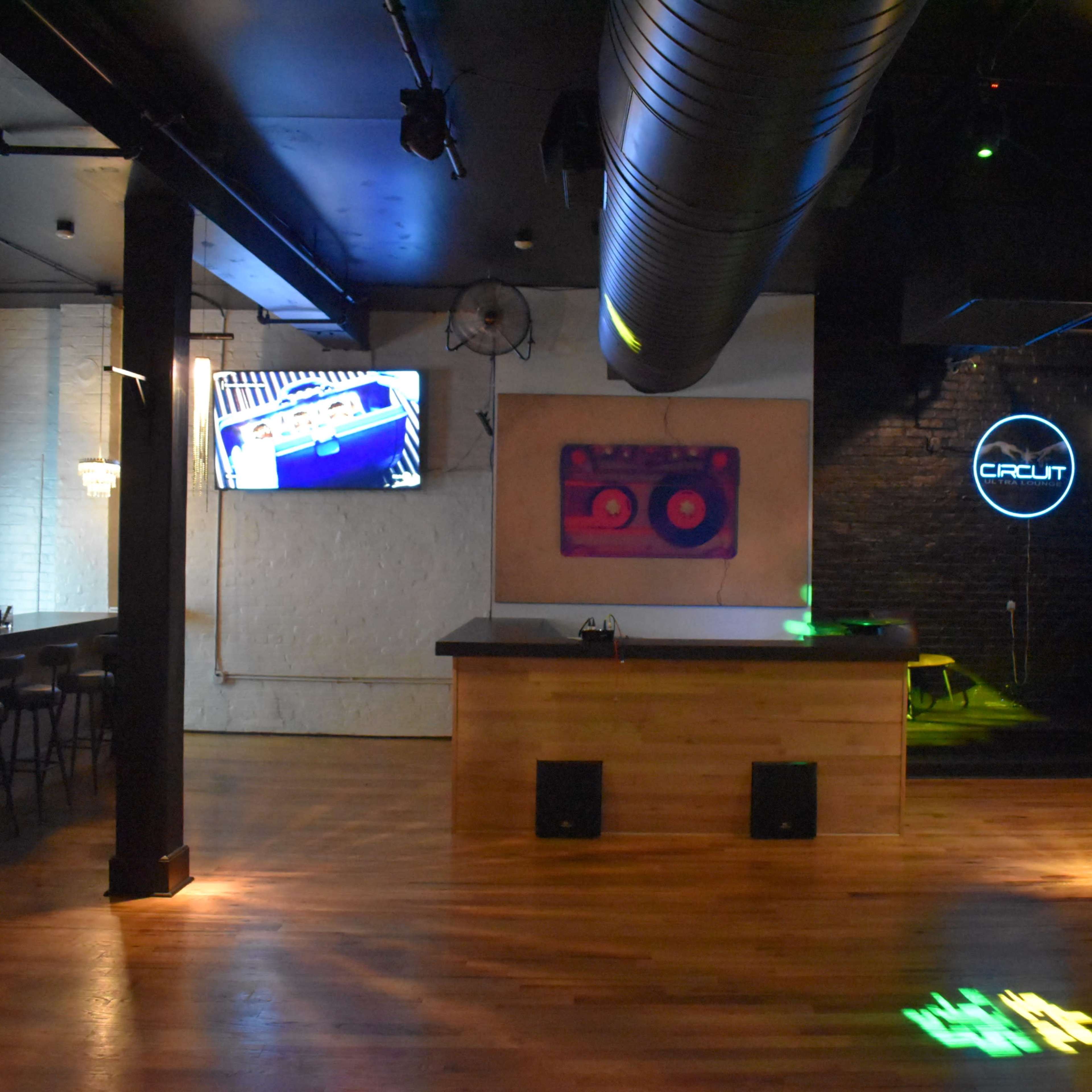 The scene depicts a modern bar setup featuring a wooden counter, high stools, and a brick wall, with screens displaying visuals and ambient lighting.