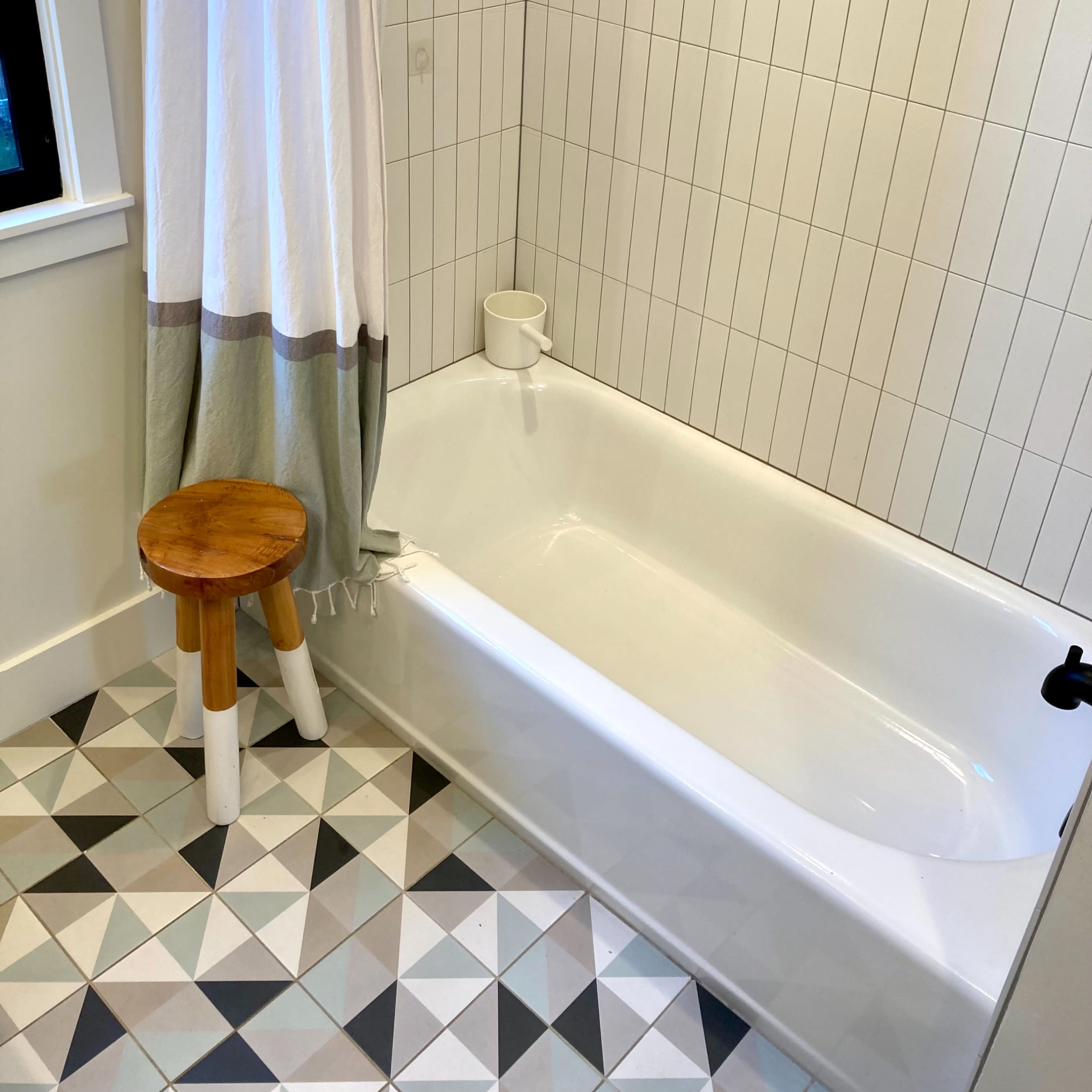 A bathroom with a white bathtub, a tiled wall, a patterned floor, and a wooden stool beside the tub.