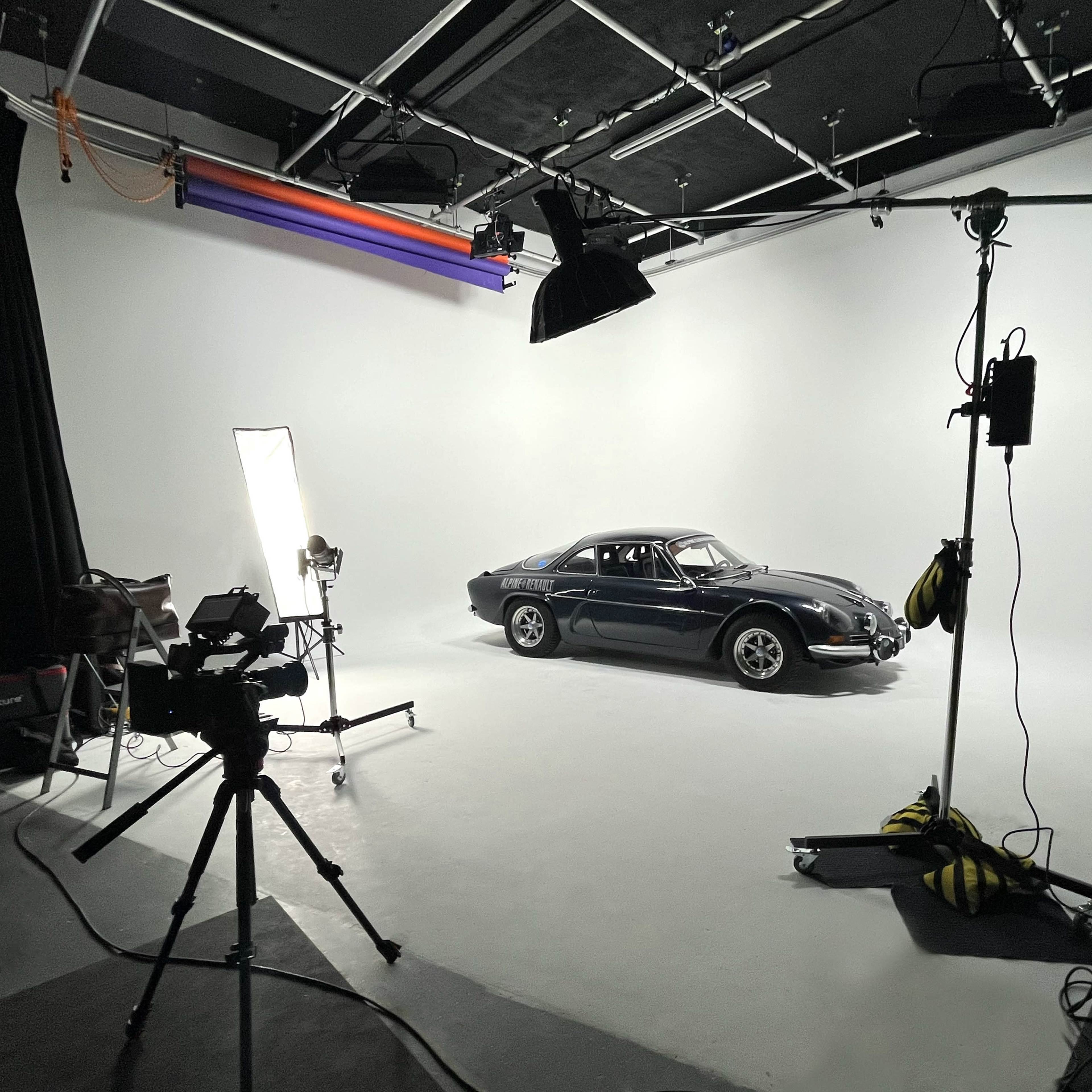 A black car is positioned on a white backdrop in a photography studio, surrounded by lighting equipment and cameras.