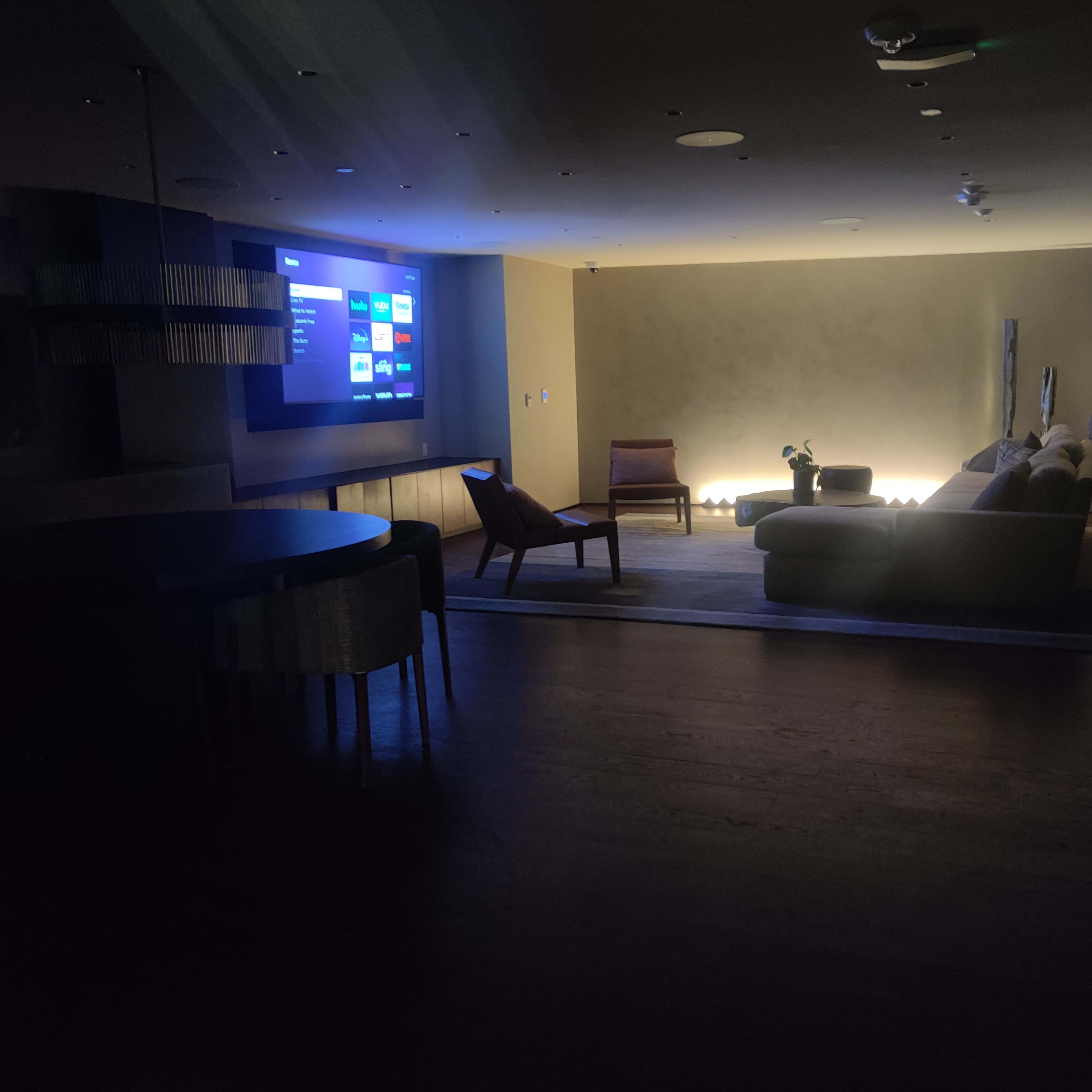 The dimly lit room features a large screen displaying content on one wall, with a sofa and a chair arranged around a low table.