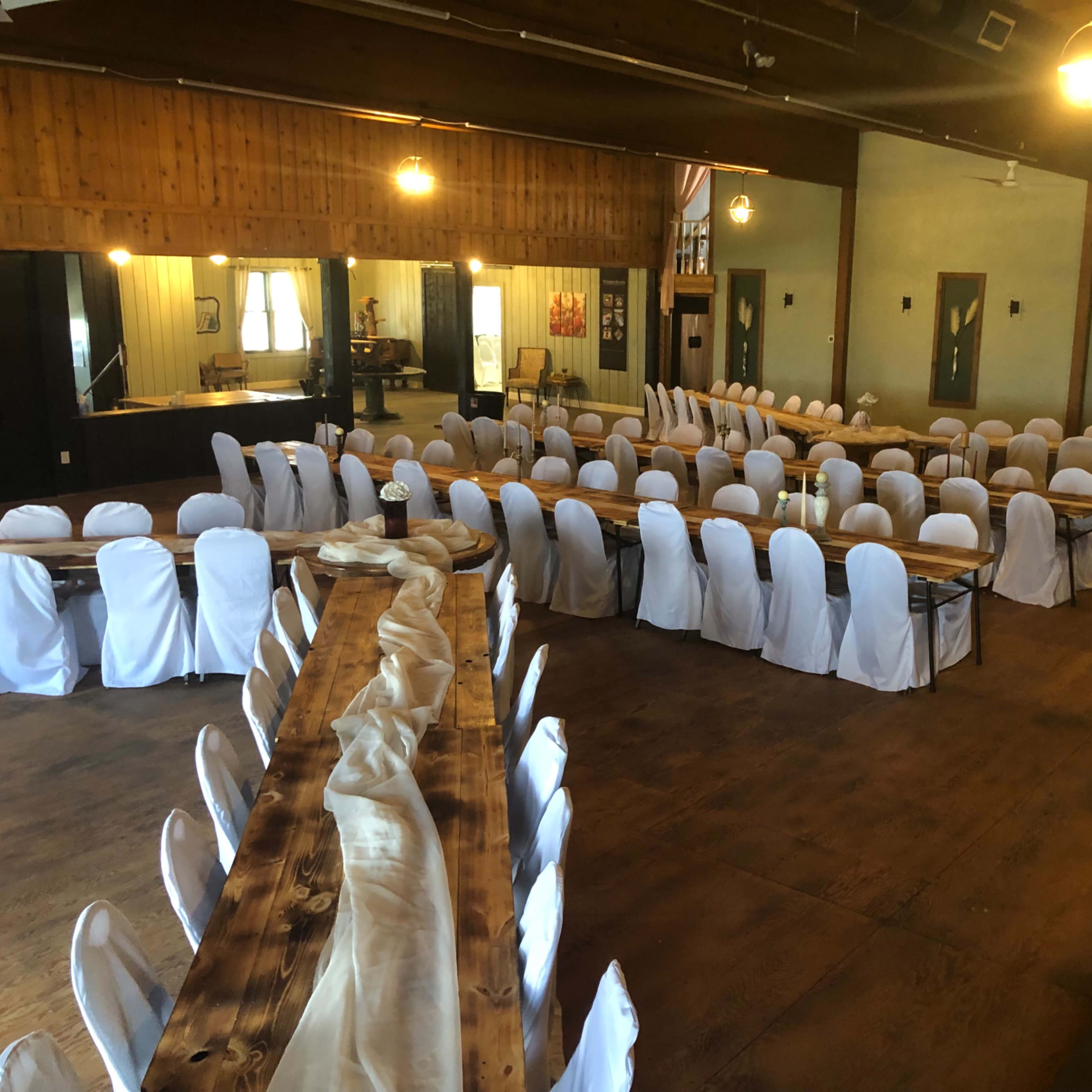 A spacious indoor venue features multiple long tables arranged with white chair covers and a central decorative table runner.