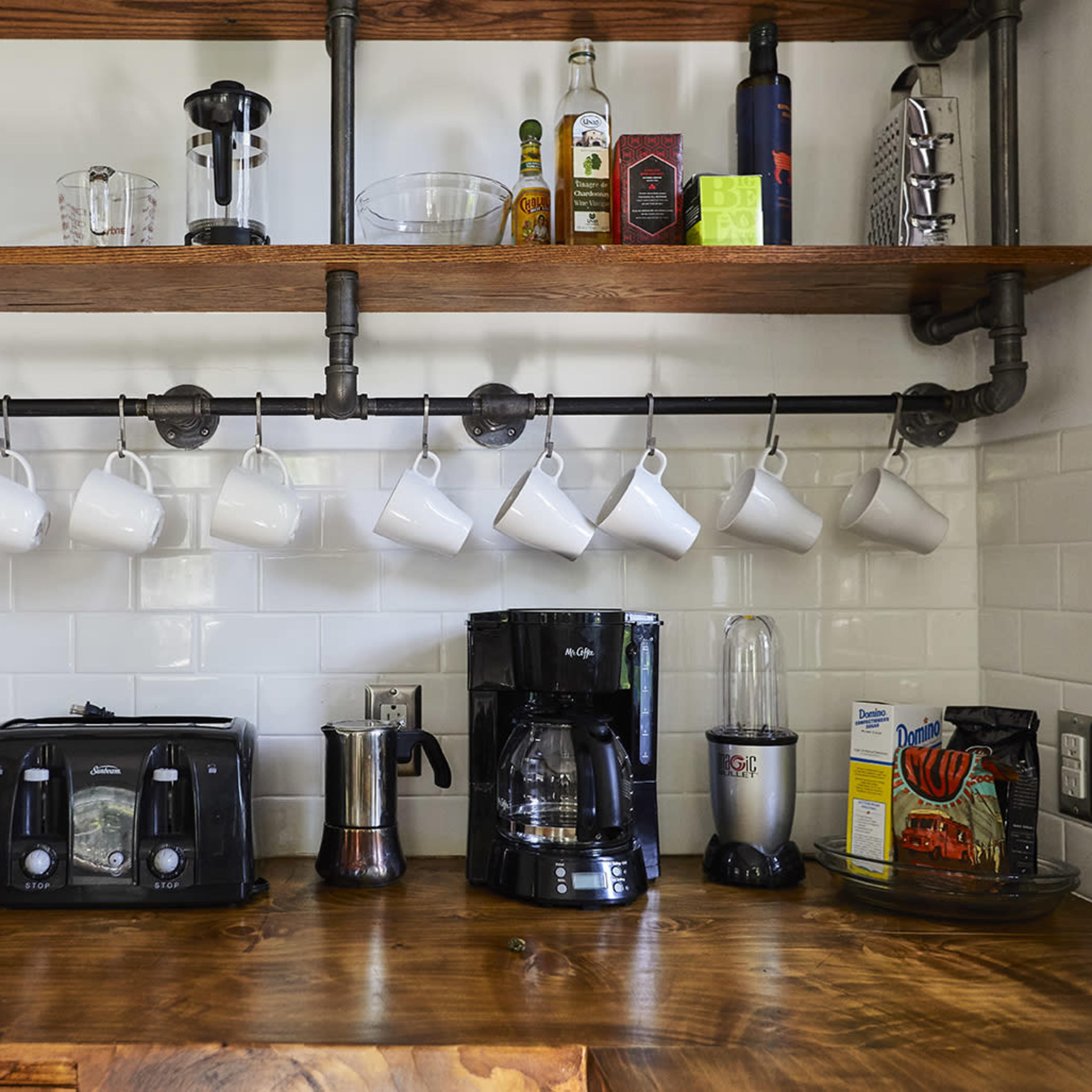 A kitchen counter displays a coffee maker, toaster, blender, and a set of hanging white mugs on a wooden shelf supported by metal pipes.