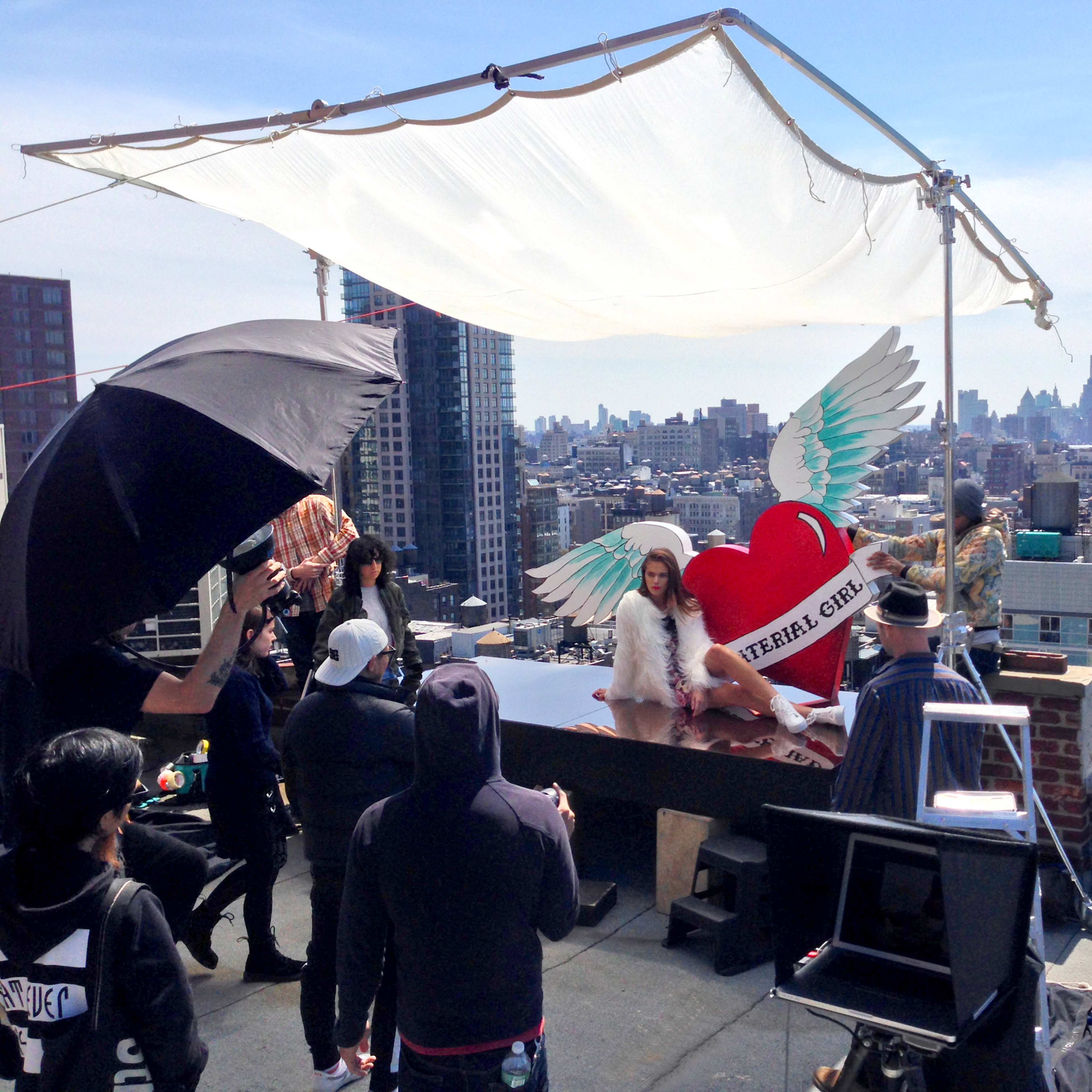 A photoshoot is taking place on a rooftop with city skyline views, featuring a model with angel wings and a large heart backdrop.